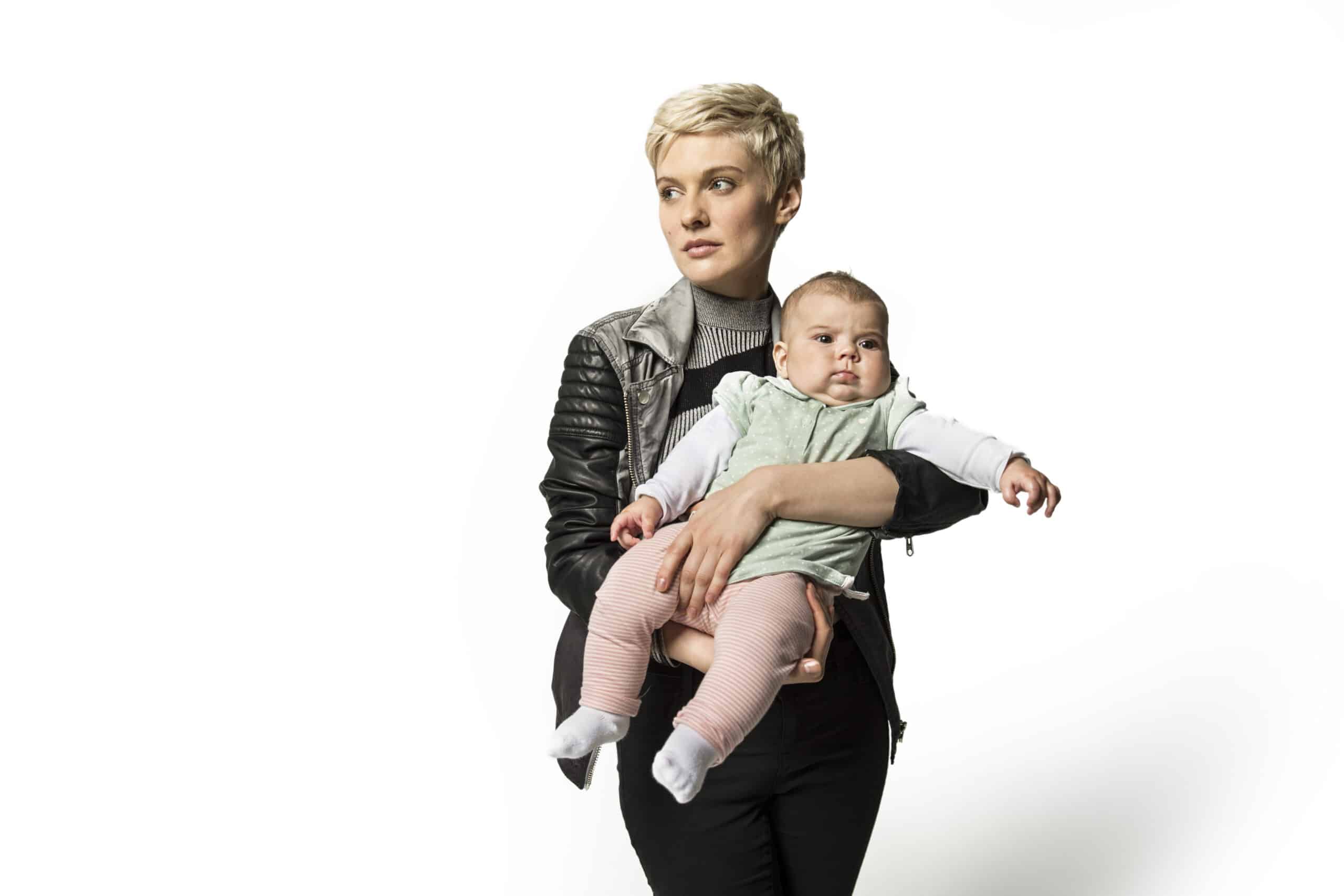 Eine Frau mit kurzen blonden Haaren, die eine schwarze Lederjacke und ein graues Oberteil trägt, hält ein Baby, das ein grünes Lätzchen und eine rosa gestreifte Hose trägt. Das Baby hat einen neugierigen Gesichtsausdruck und streckt einen Arm aus. Der Hintergrund ist schlicht weiß. © Fotografie Tomas Rodriguez