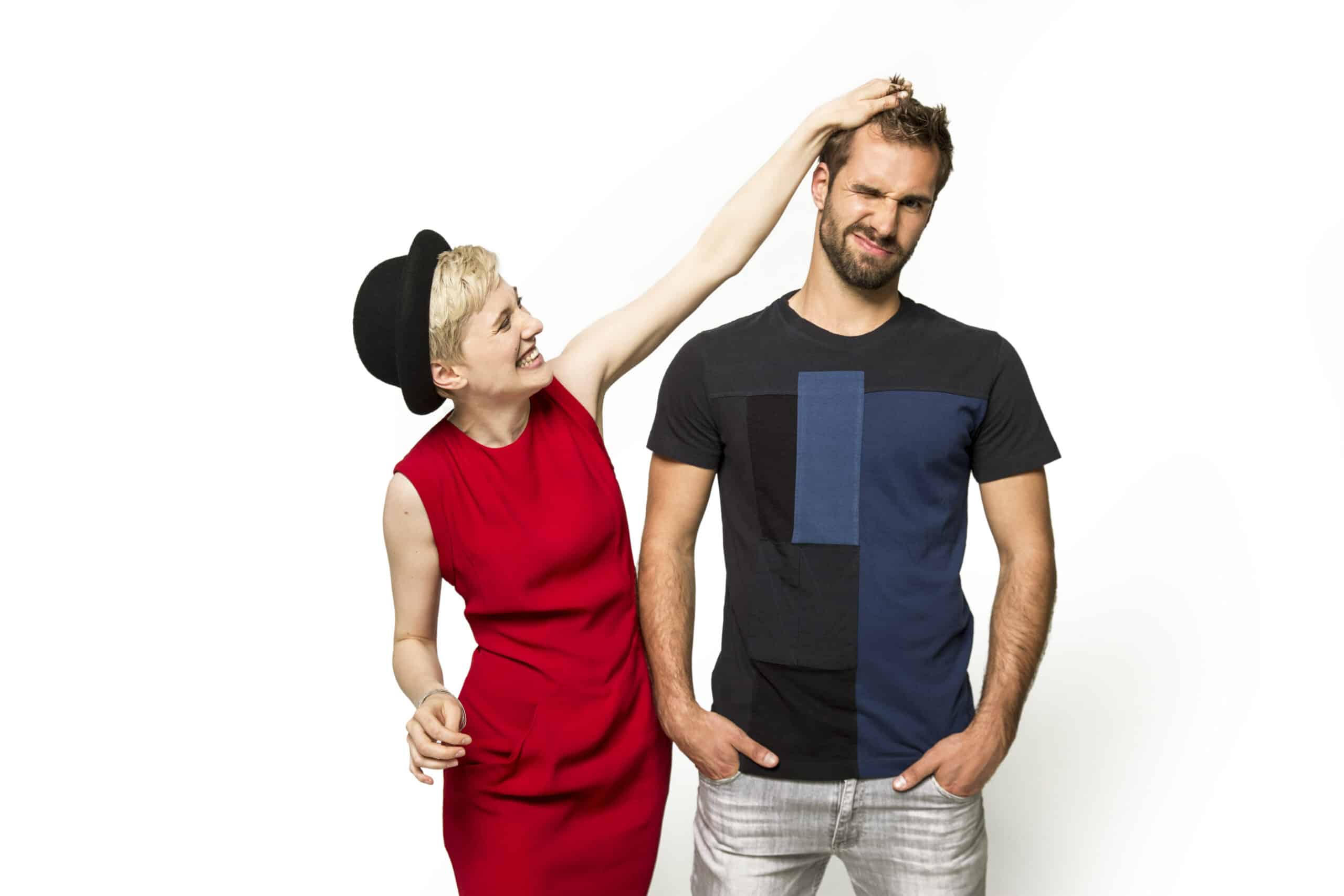 Eine lächelnde Frau in einem roten Kleid und einem schwarzen Hut zieht spielerisch an den Haaren eines Mannes in einem schwarz-blauen T-Shirt mit verzogenem Gesicht. Der Mann steht mit den Händen in den Taschen und beide stehen vor einem schlichten weißen Hintergrund. © Fotografie Tomas Rodriguez