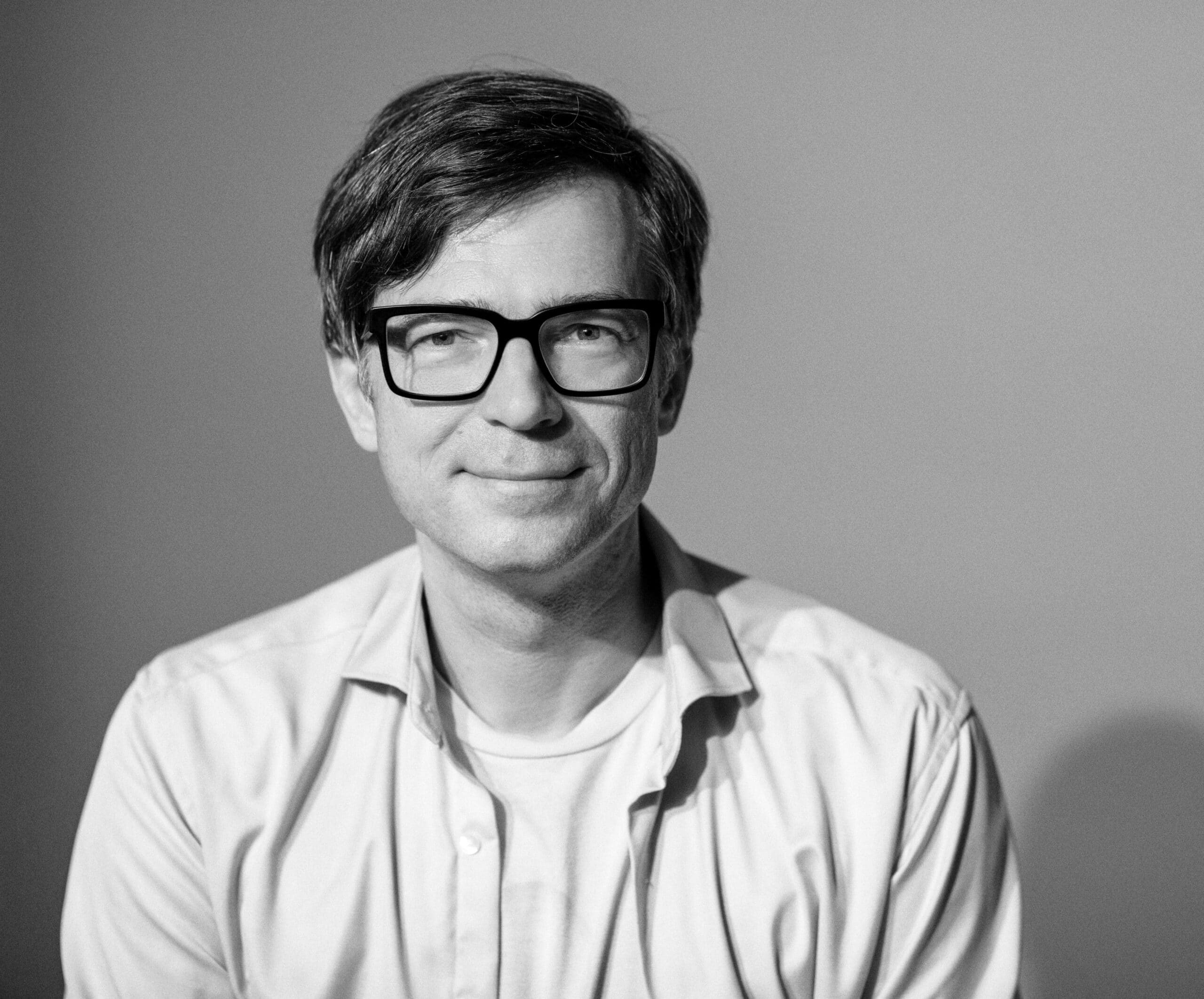 Schwarzweißporträt eines Mannes mit Brille und Button-Down-Hemd. Er hat kurze Haare und lächelt vor einem neutralen Hintergrund. © Fotografie Tomas Rodriguez