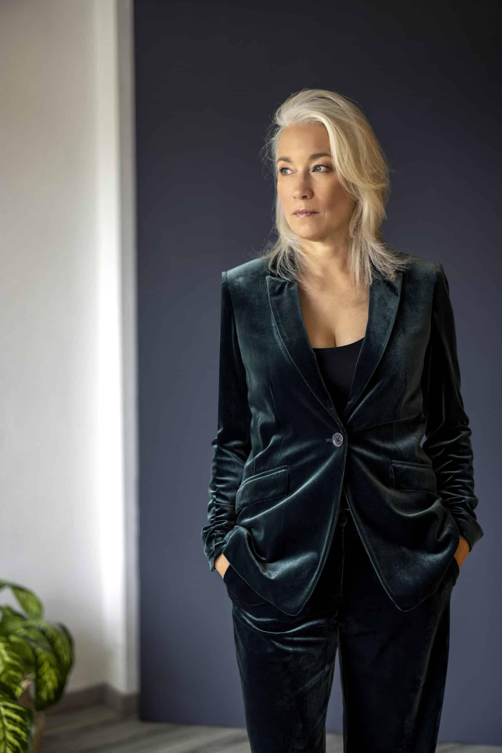 Eine Frau mit platinblondem Haar steht selbstbewusst da. Sie trägt ein dunkelgrünes Samtkostüm und hat die Hände in den Taschen. Sie blickt mit nachdenklichem Gesichtsausdruck zur Seite. Der Hintergrund ist eine einfache, dunkel gefärbte Wand, in deren Ecke eine kleine Pflanze zu sehen ist. © Fotografie Tomas Rodriguez