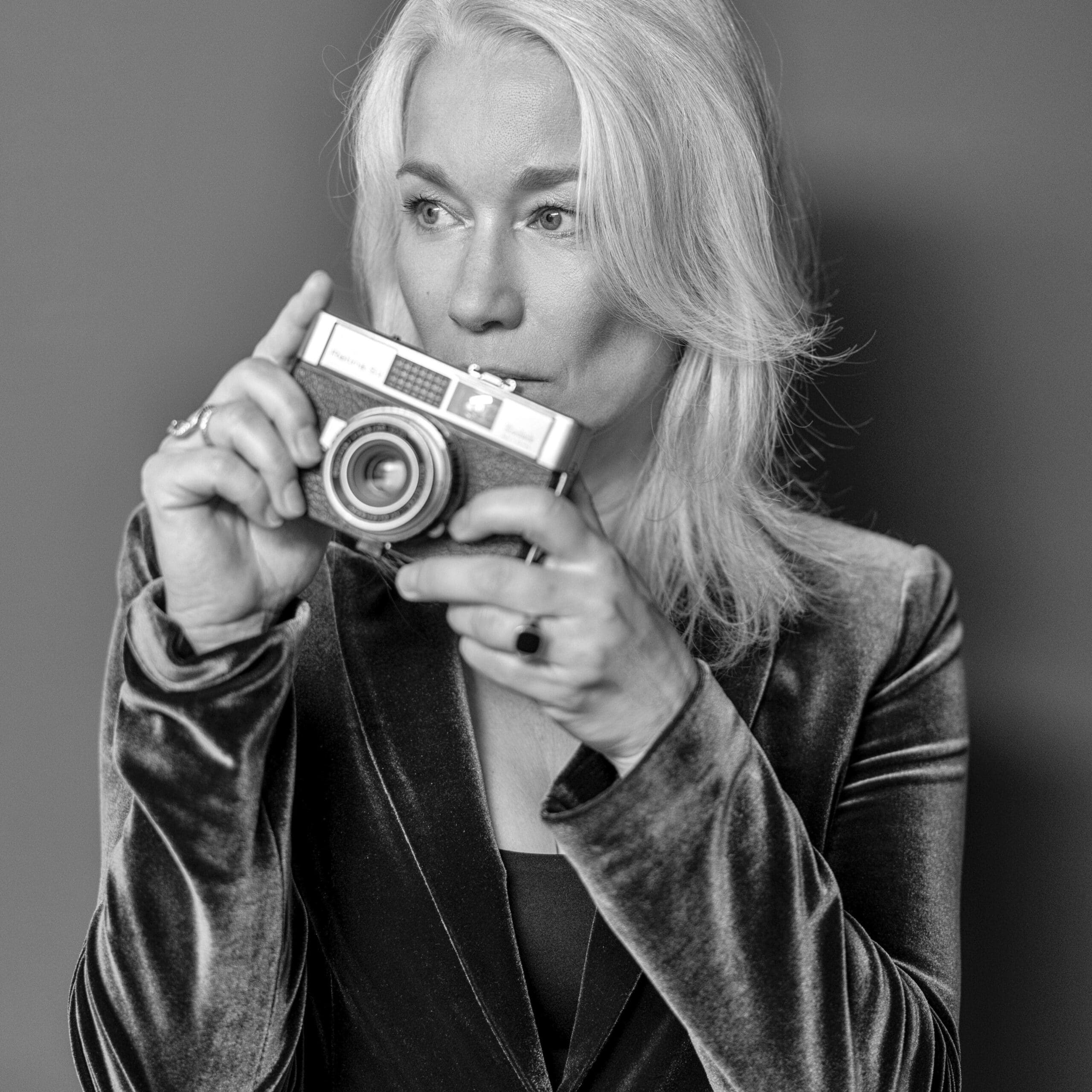 Schwarzweißbild einer Person mit langem, hellem Haar, die eine Vintage-Kamera vor ihr Gesicht hält. Die Person trägt eine Samtjacke und blickt aufmerksam zur Seite, während sie die Kamera einstellt. Der Hintergrund ist schlicht. © Fotografie Tomas Rodriguez
