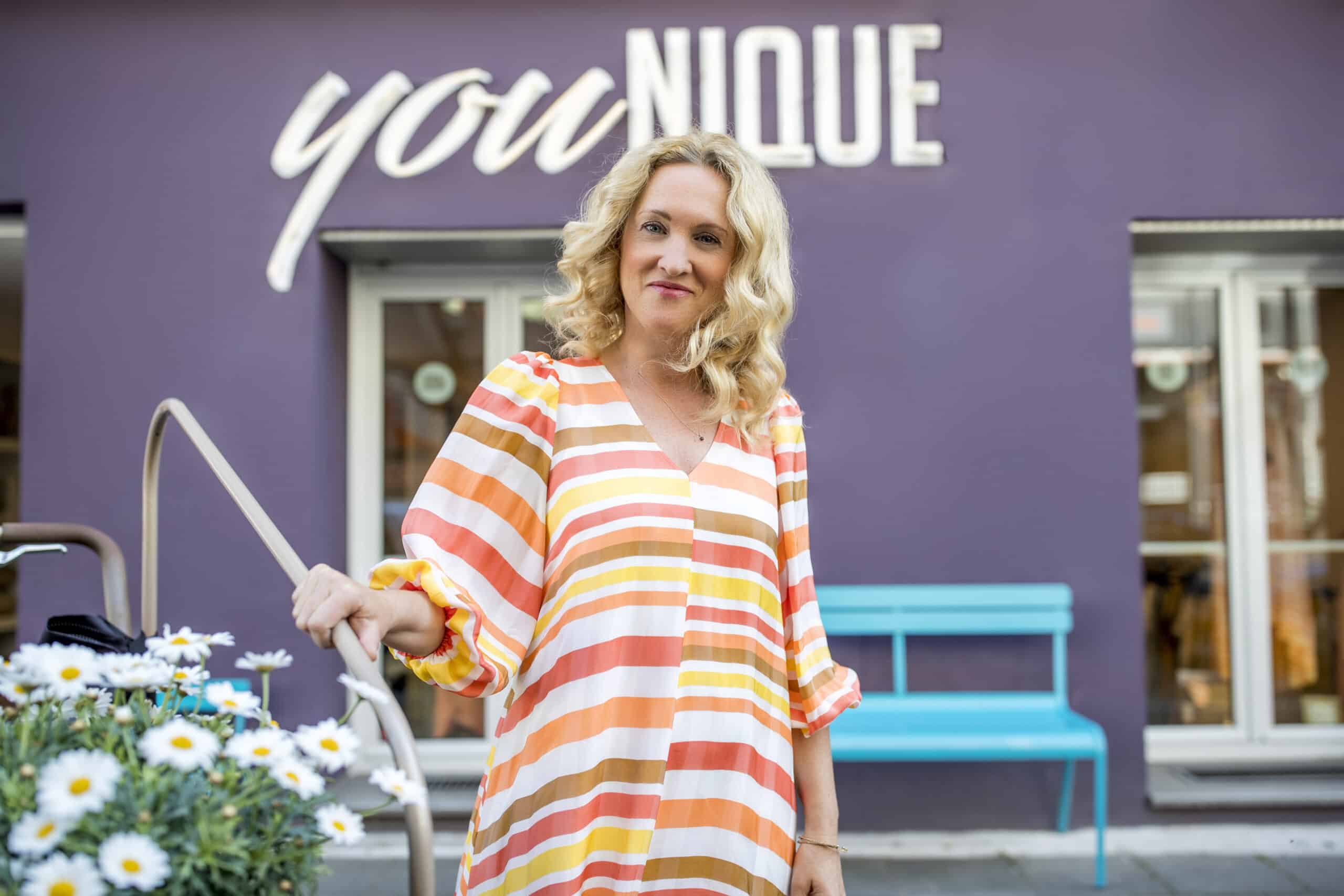 Eine Frau mit blonden Haaren und einem bunt gestreiften Kleid steht vor einem Gebäude, hinter dem das Wort „YOUnique“ prangt. Sie hält ein Geländer neben einer Topfpflanze mit weißen Blüten. Im Hintergrund sind eine blaue Bank und Fenster zu sehen. © Fotografie Tomas Rodriguez