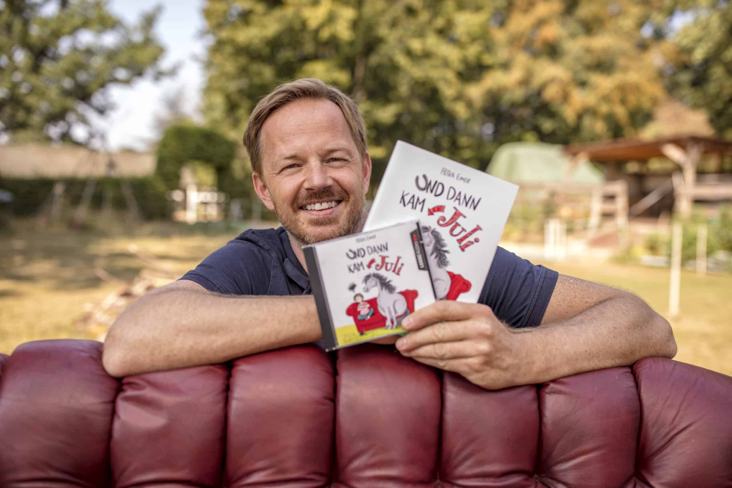 Ein Mann mit hellem Bart und kurzen Haaren beugt sich lächelnd über die Rückenlehne eines roten Sofas und hält Bücher mit dem Titel „Und dann kam Juli!“ hoch. Eines davon ist ein Buch in CD-Größe, das andere eine Ausgabe in Buchgröße. Im Hintergrund ist ein Garten mit Bäumen und Gartenmöbeln zu sehen. © Fotografie Tomas Rodriguez