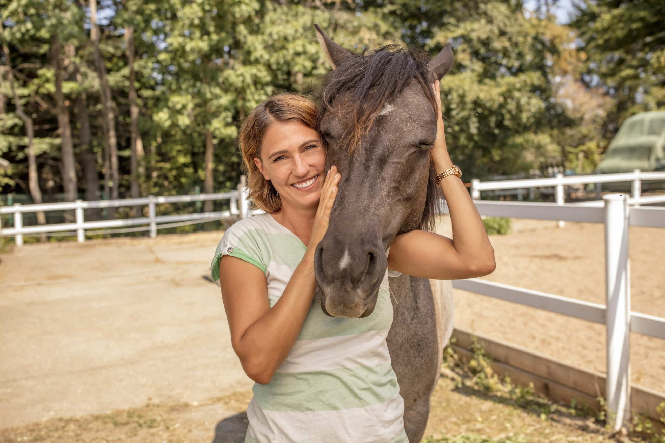 Eine lächelnde Frau steht in einem Pferdegehege und hält liebevoll den Kopf eines grauen Pferdes. Beide sind von einem weißen Zaun umgeben und im Hintergrund sind Bäume zu sehen. Die Frau trägt ein helles, gestreiftes Hemd. © Fotografie Tomas Rodriguez
