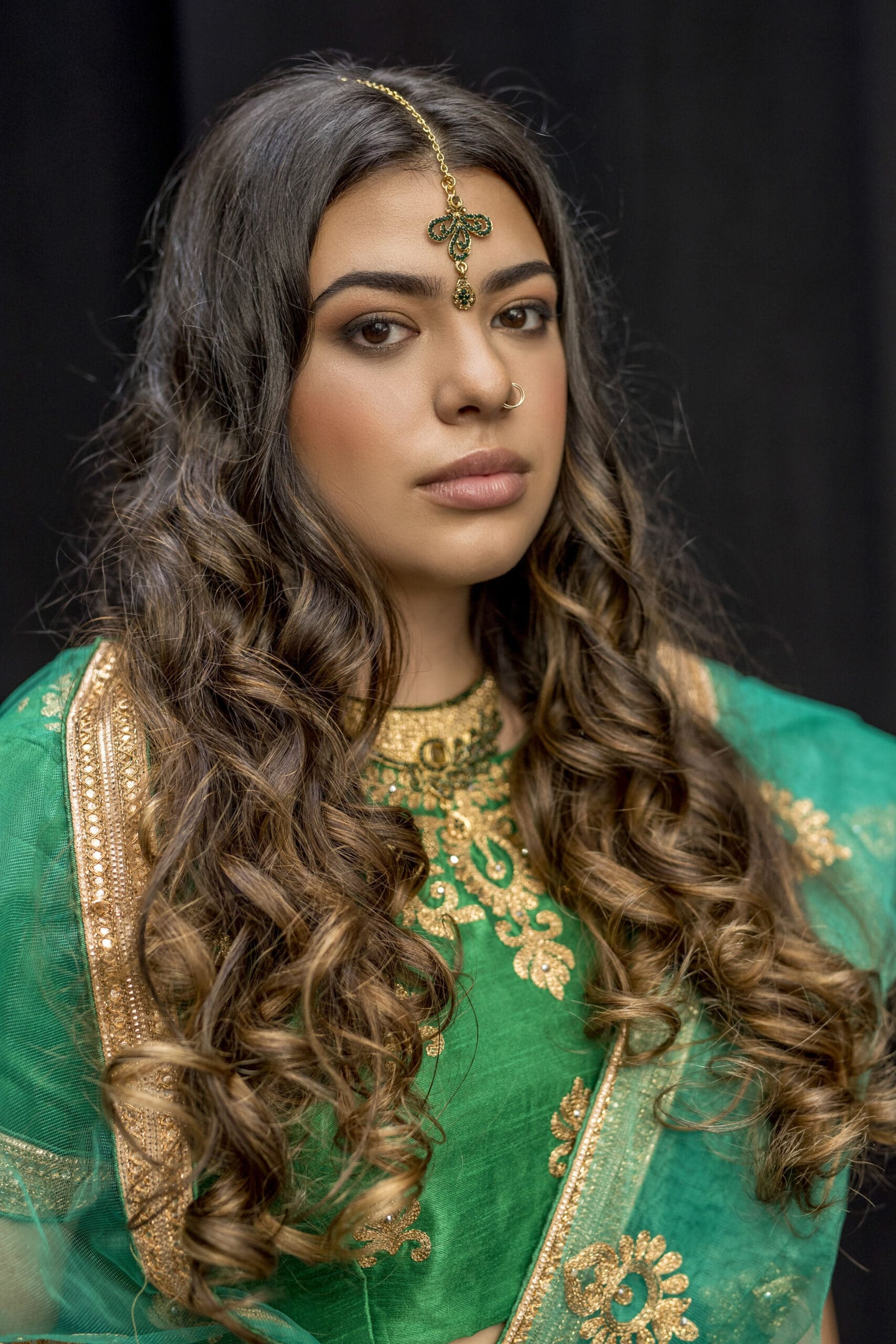 Eine Frau mit langem, welligem Haar trägt ein grünes, traditionelles Outfit, das mit Goldstickereien verziert ist. Sie hat einen Nasenring und ein dekoratives Kopfstück mit einer Kette und einem Ornament, das an ihrer Stirn hängt. Der Hintergrund ist schwarz, wodurch sie zum Mittelpunkt des Bildes wird. © Fotografie Tomas Rodriguez