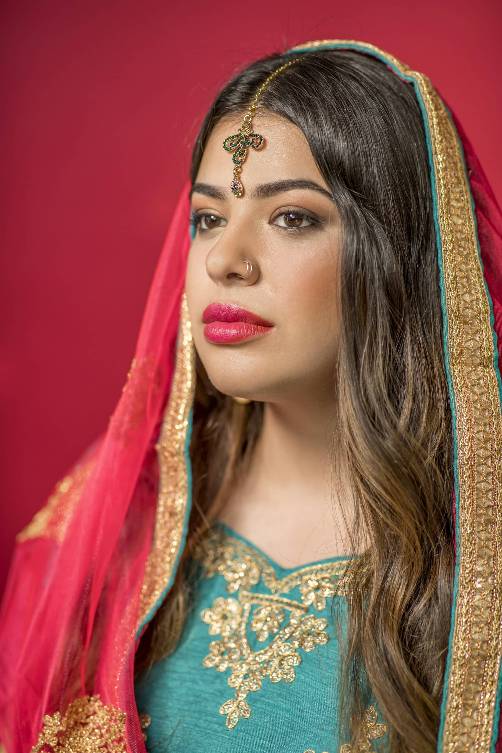 Eine Frau mit langem, dunklem Haar trägt ein traditionelles Outfit. Sie hat einen dekorativen Maang Tikka auf der Stirn, einen Nasenring und einen leuchtend roten und goldenen Dupatta über dem Kopf. Der Hintergrund ist ein sattes Rot, das ihre Kleidung ergänzt. © Fotografie Tomas Rodriguez