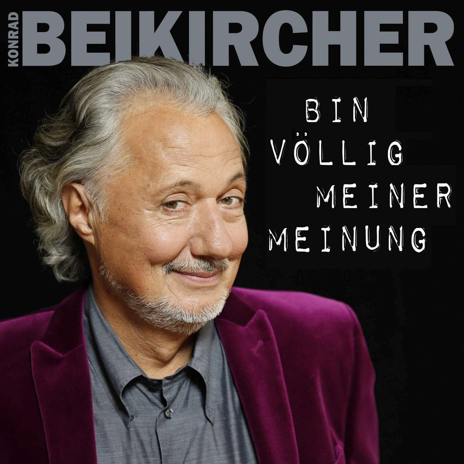 Ein Mann mit grauem Haar und Bart grinst leicht in die Kamera. Er trägt einen lila Samtblazer und ein graues Hemd. Der Hintergrund ist dunkel, oben steht der Name „Konrad Beikircher“ und rechts der Text „Bin völlig meiner Meinung“. © Fotografie Tomas Rodriguez
