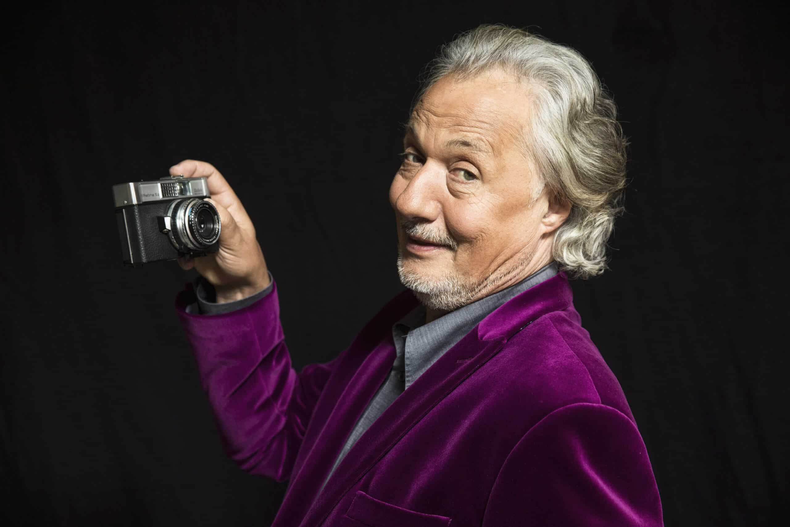 Ein älterer Mann mit welligem weißem Haar und kurzem Bart trägt eine leuchtend violette Samtjacke und hält mit der rechten Hand eine Vintage-Kamera hoch. Er lächelt selbstbewusst und dreht seinen Kopf leicht nach links. Der Hintergrund ist schwarz. © Fotografie Tomas Rodriguez