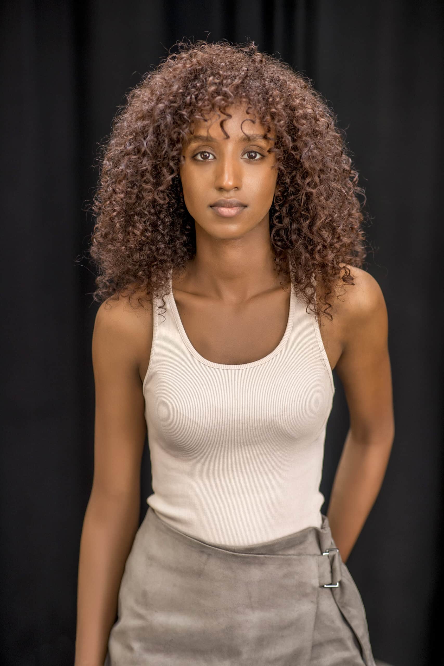 Eine Frau mit lockigem braunem Haar trägt ein beiges Tanktop und einen grauen Rock mit seitlichen Trägern. Sie steht vor einem schwarzen Hintergrund und blickt mit gelassenem Gesichtsausdruck in die Kamera. © Fotografie Tomas Rodriguez
