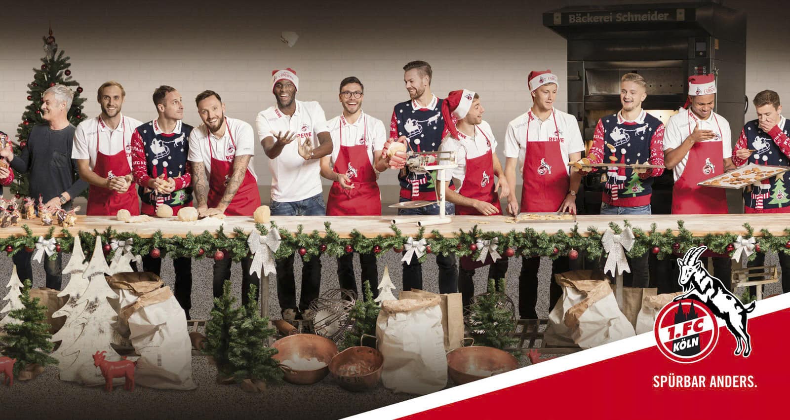 Eine Gruppe von Menschen, darunter Spieler und Mitarbeiter des 1. FC Köln, steht hinter einem langen Tisch, der mit festlichen Girlanden geschmückt und mit Backwaren gefüllt ist. Sie tragen rote Schürzen und einige haben Weihnachtsmannmützen auf. Hinter ihnen hängt ein Schild mit der Aufschrift „Bäckerei Schneider“. © Fotografie Tomas Rodriguez