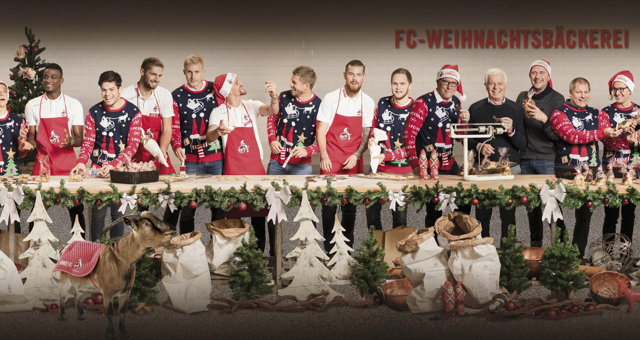 Eine Gruppe von Menschen, viele davon tragen passende Weihnachtspullover und rote Schürzen, steht hinter einem langen, festlichen Tisch, der mit Girlanden und Ornamenten geschmückt ist. Auf dem Tisch sind verschiedene Backwaren ausgestellt, und in der Szene sind ein Weihnachtsbaum und eine Ziege zu sehen. Auf dem Schild steht „FC-WEIHNACHTSBÄCKEREI“. © Fotografie Tomas Rodriguez