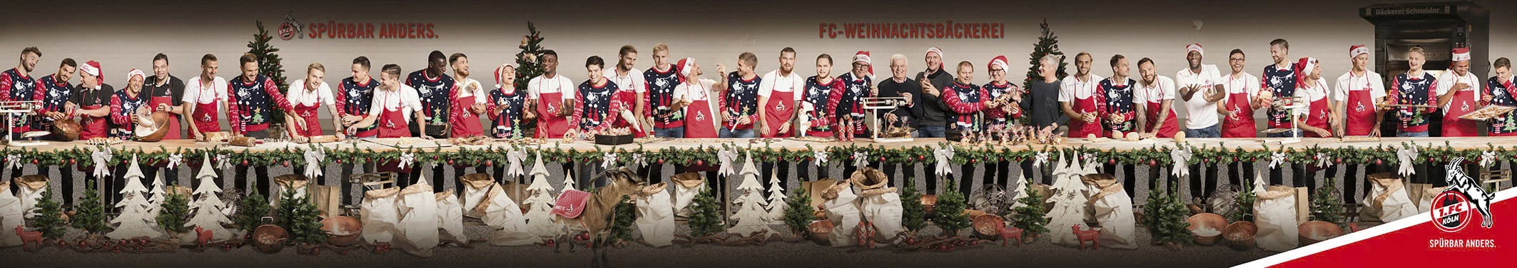 An einem langen Tisch, der mit Girlanden, Tannenzapfen und kleinen Weihnachtsbäumen geschmückt ist, sitzen 32 Personen in festlichen, weihnachtlichen Schürzen und Weihnachtsmannmützen. Sie sind mit verschiedenen Kochaktivitäten beschäftigt, vom Schneiden der Zutaten bis zum Mischen und Präsentieren der Gerichte. © Fotografie Tomas Rodriguez
