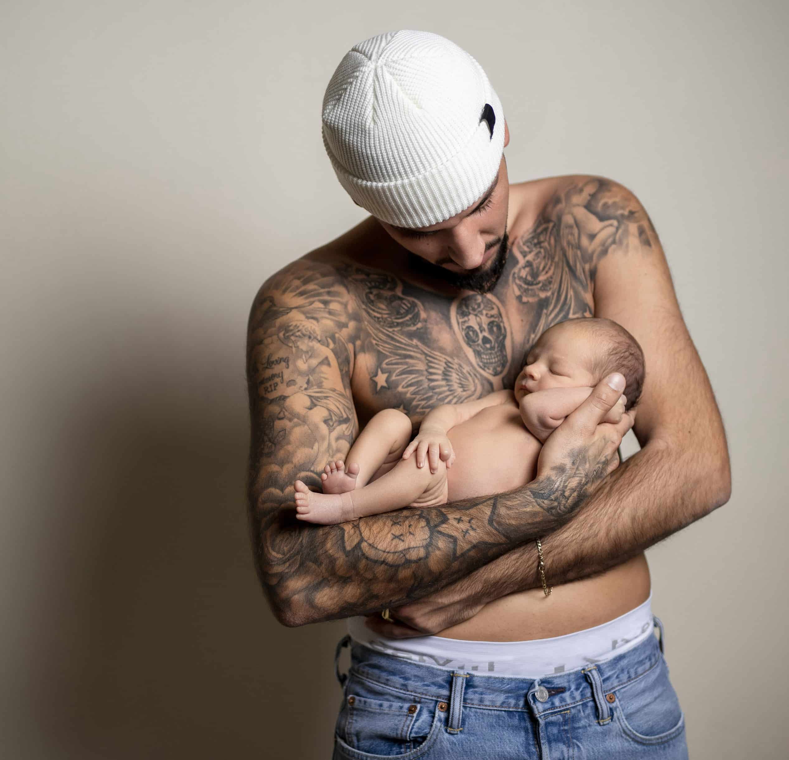 Ein Mann ohne Hemd mit Tätowierungen auf Oberkörper und Armen wiegt ein schlafendes Neugeborenes in seinen Armen. Der Mann trägt eine weiße Mütze und Jeans und blickt mit zärtlichem Gesichtsausdruck auf das Baby herab. Der Hintergrund ist in einer schlichten neutralen Farbe gehalten. © Fotografie Tomas Rodriguez