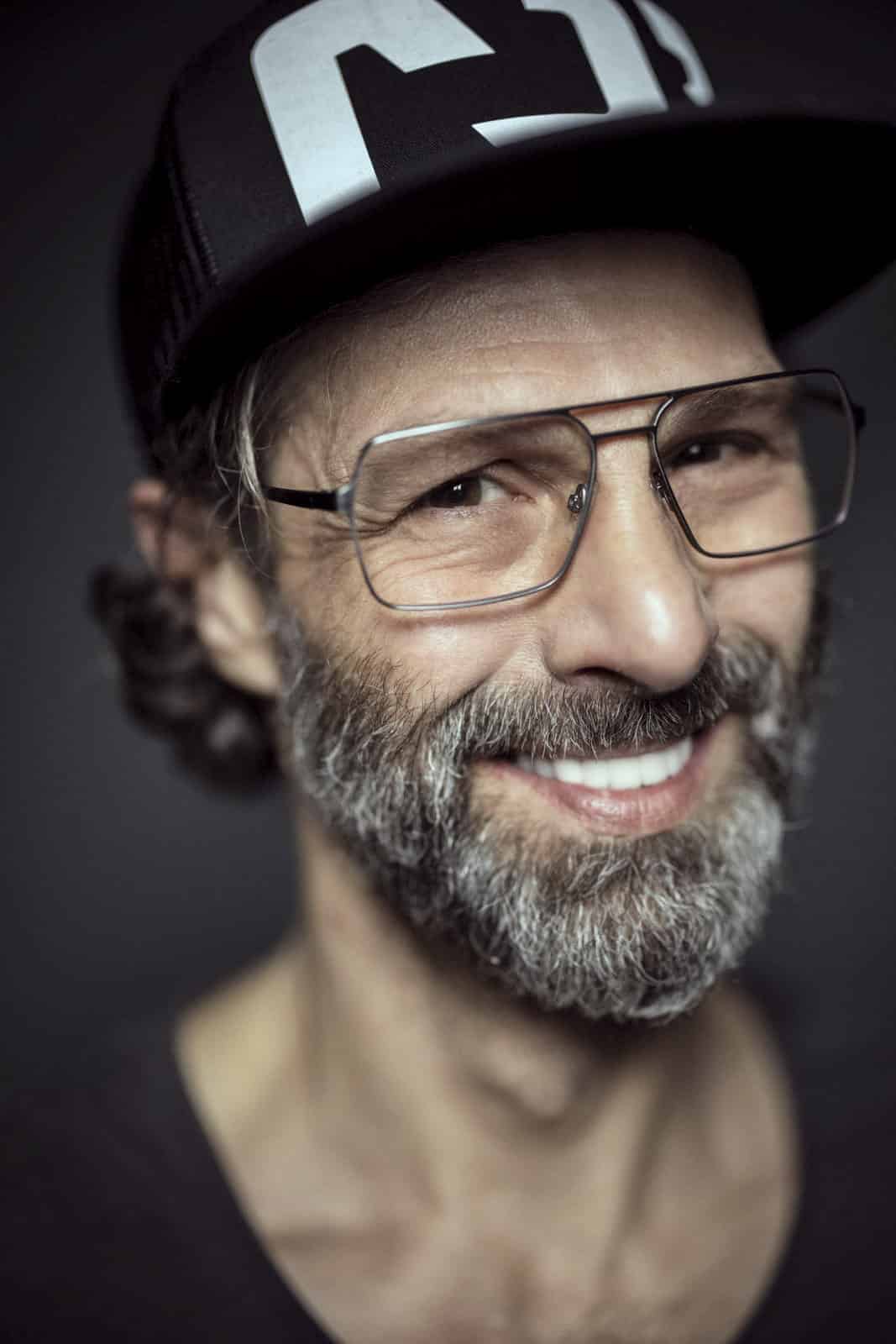 Ein Mann mit grauem Bart und Schnurrbart lächelt in die Kamera. Er trägt eine große, rechteckige Brille und eine schwarze Baseballkappe mit weißen Buchstaben auf der Vorderseite. Der Hintergrund ist dunkel und hebt sein Gesicht hervor. © Fotografie Tomas Rodriguez