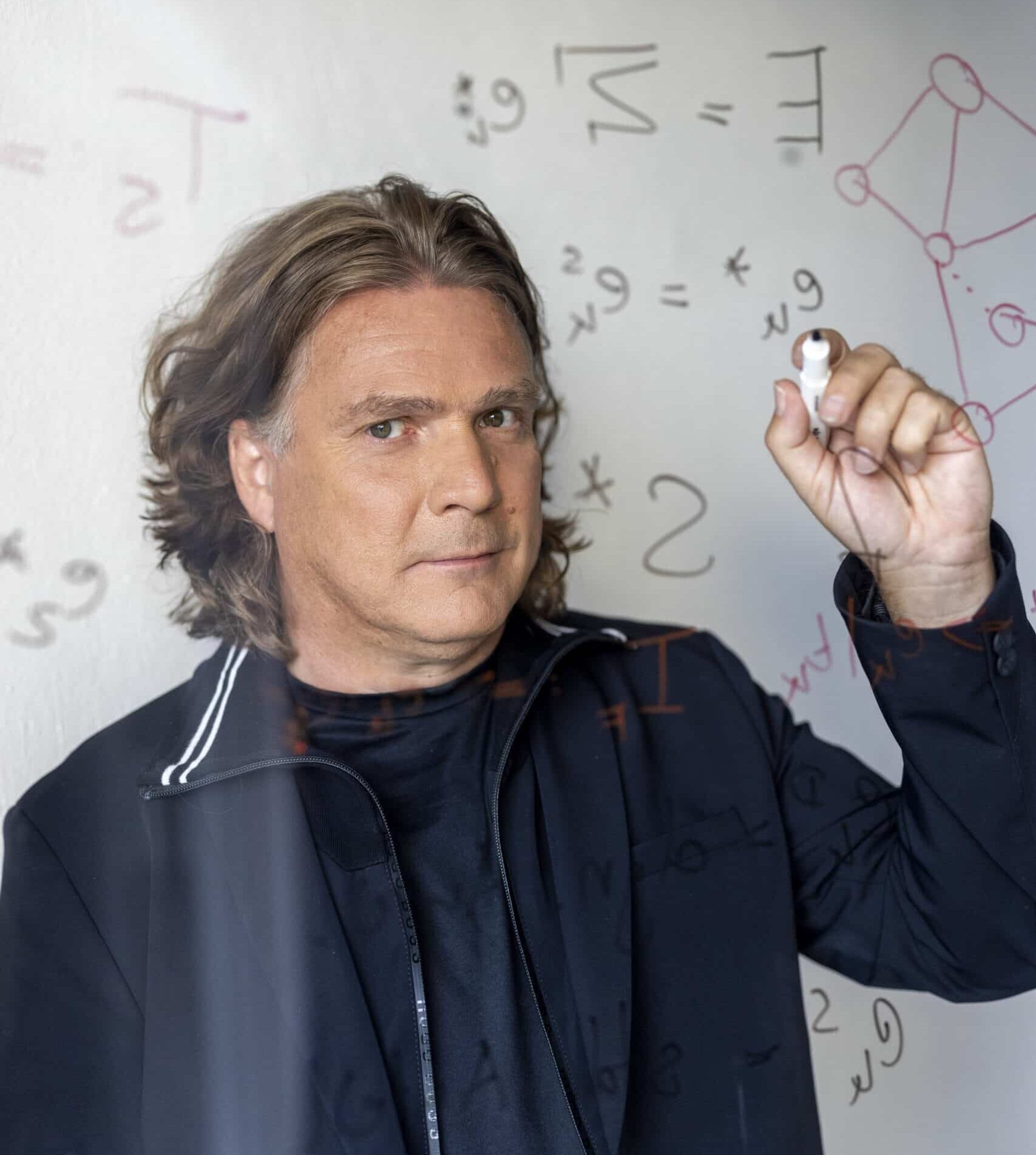 Eine Person mit schulterlangem Haar schreibt mit einem Marker mathematische Gleichungen auf eine Glastafel. Sie trägt eine dunkle Jacke und blickt aufmerksam auf die Tafel, auf deren Glas sich die Gleichungen spiegeln. © Fotografie Tomas Rodriguez
