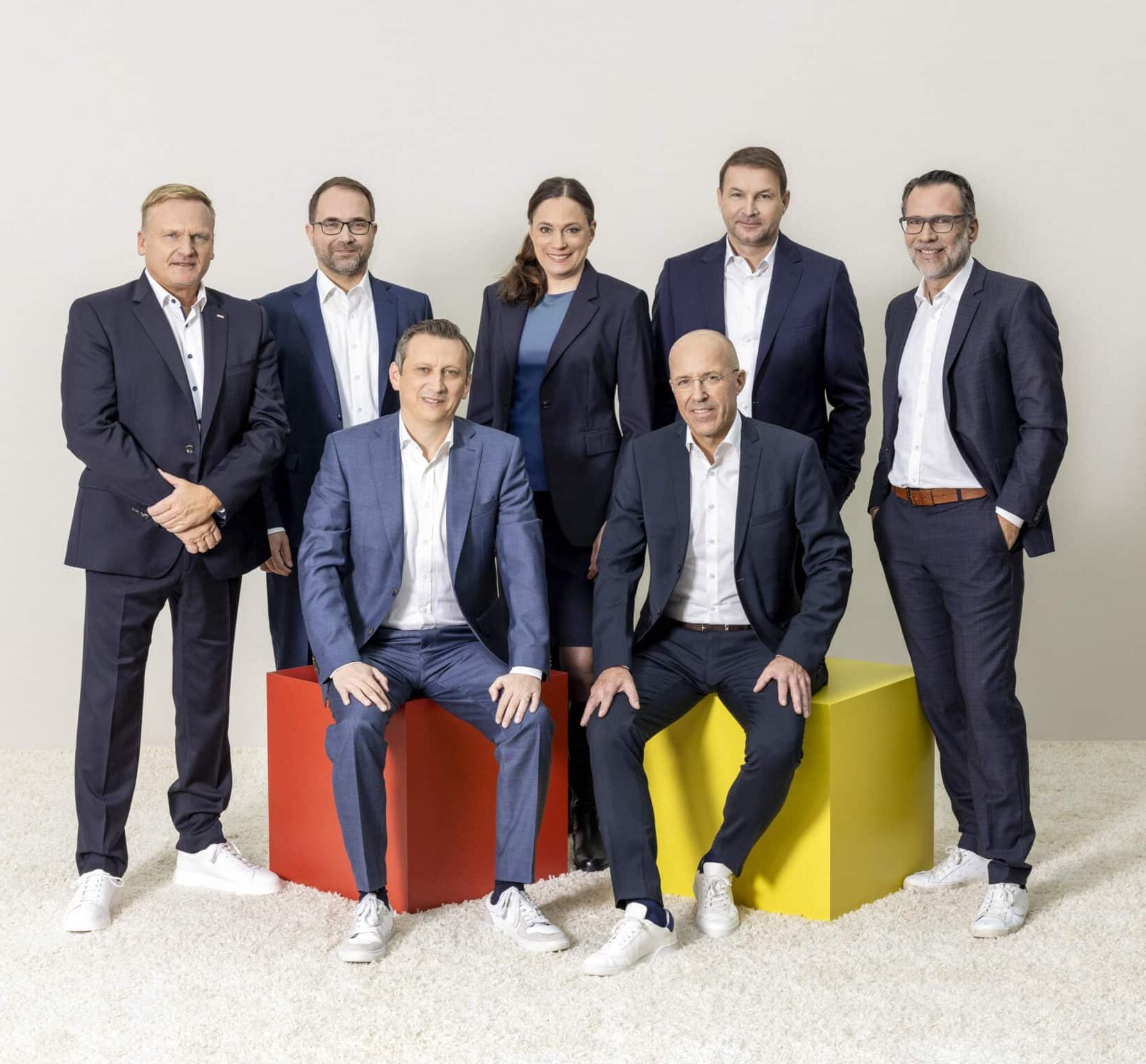 Eine Gruppe von sieben professionell gekleideten Personen, bestehend aus sechs Männern und einer Frau, posiert für ein formelles Foto. Zwei Männer sitzen auf bunten Sitzwürfeln (einer rot, einer gelb), während die anderen hinter ihnen vor einem schlichten hellen Hintergrund stehen. © Fotografie Tomas Rodriguez