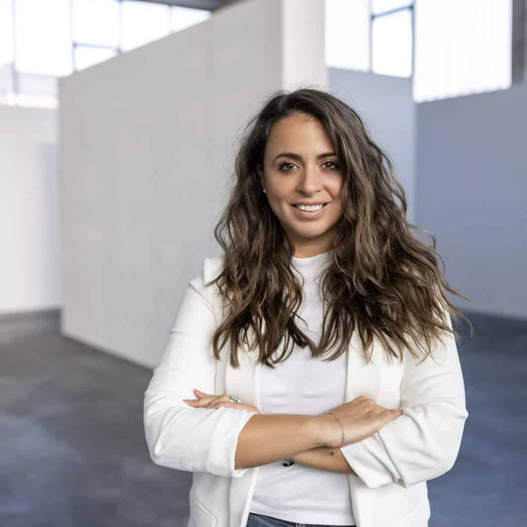 Eine Frau mit langen, welligen Haaren und einem weißen Blazer steht mit verschränkten Armen in einem modernen Raum im Industriestil mit weißen Wänden und großen Fenstern. Sie lächelt und wirkt selbstbewusst. © Fotografie Tomas Rodriguez