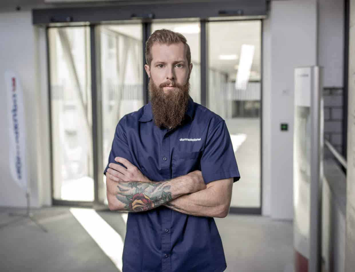 Ein Mann mit langem Bart und Tattoos auf dem rechten Arm steht mit verschränkten Armen da. Er trägt ein marineblaues Kurzarmhemd und befindet sich in einem modernen, gut beleuchteten Raum mit großen Fenstern im Hintergrund. © Fotografie Tomas Rodriguez