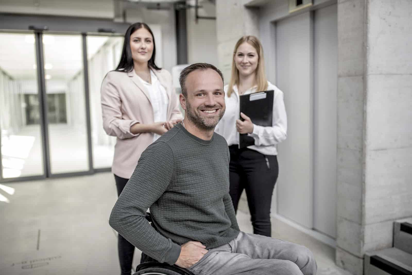 Im Vordergrund steht ein lächelnder Mann im Rollstuhl, hinter ihm stehen zwei Frauen. Die Frau links hält die Griffe des Rollstuhls, die Frau rechts hält einen schwarzen Ordner. Sie befinden sich in einem modernen, hellen Flur mit Glastüren. © Fotografie Tomas Rodriguez