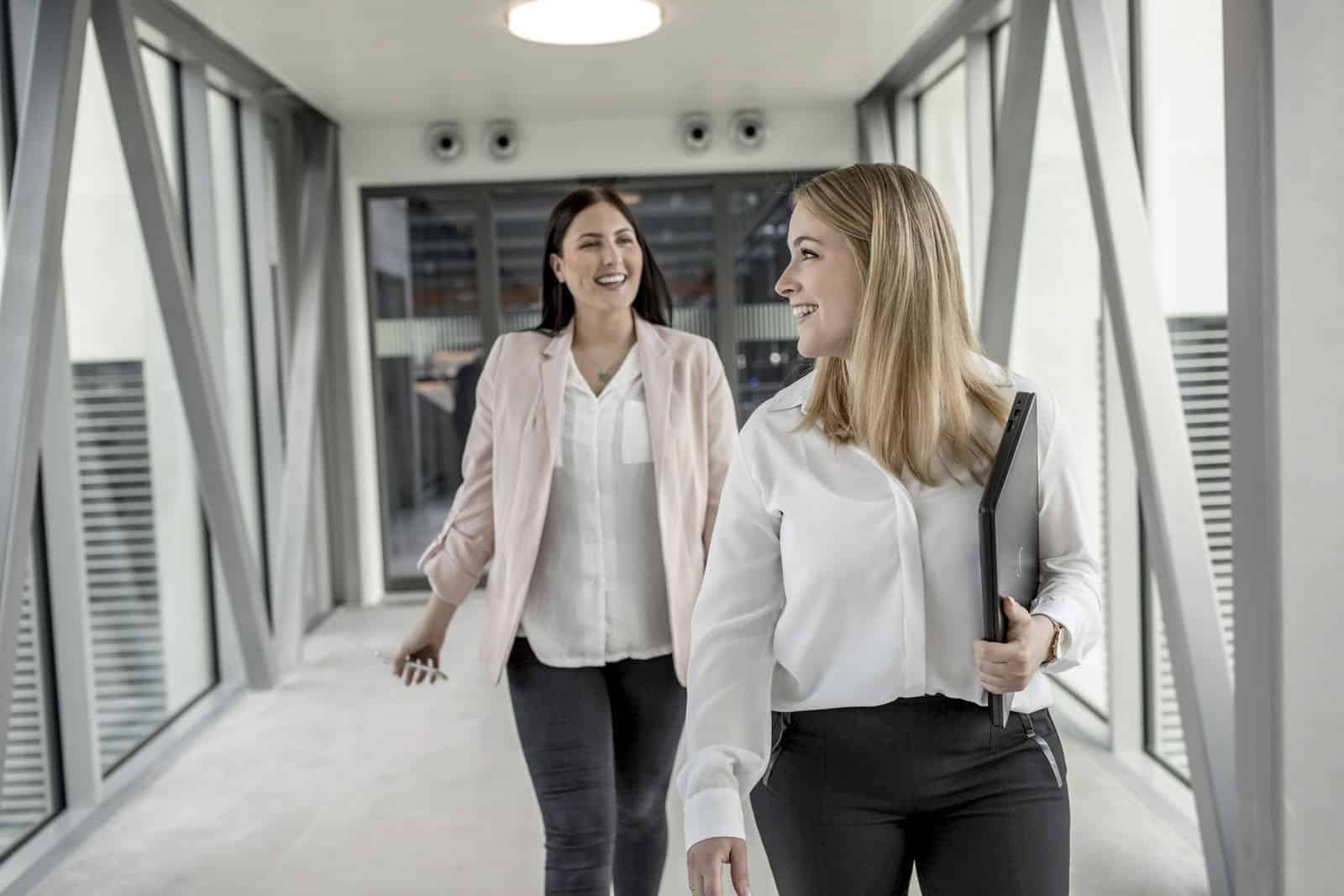 Zwei Frauen gehen durch einen modernen Büroflur. Beide lächeln und sind leger gekleidet. Die Frau im Vordergrund hält einen Ordner in der Hand. Im Hintergrund sind Glaswände und ein Metallgerüst zu sehen, was an eine moderne Büroumgebung erinnert. © Fotografie Tomas Rodriguez