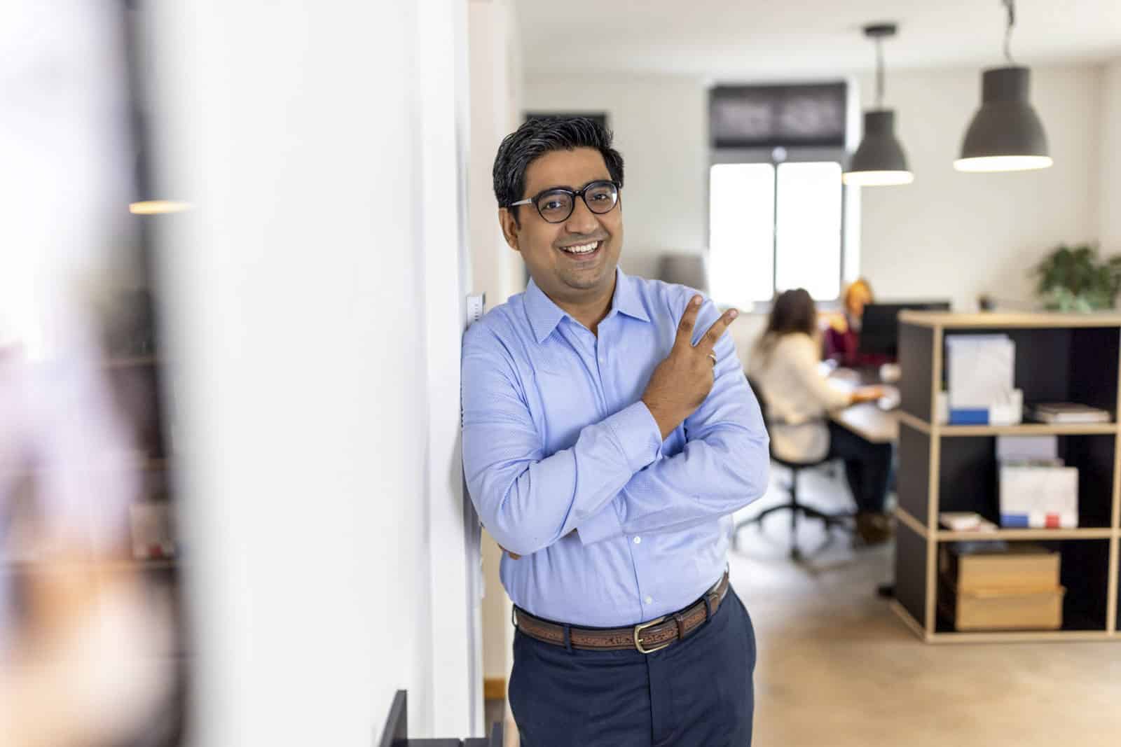 Ein lächelnder Mann mit Brille und blauem Hemd steht mit verschränkten Armen an einer Wand gelehnt und macht mit seiner rechten Hand ein Peace-Zeichen. Im Hintergrund arbeiten zwei Personen an Schreibtischen in einem gut beleuchteten, modernen Büroraum. © Fotografie Tomas Rodriguez