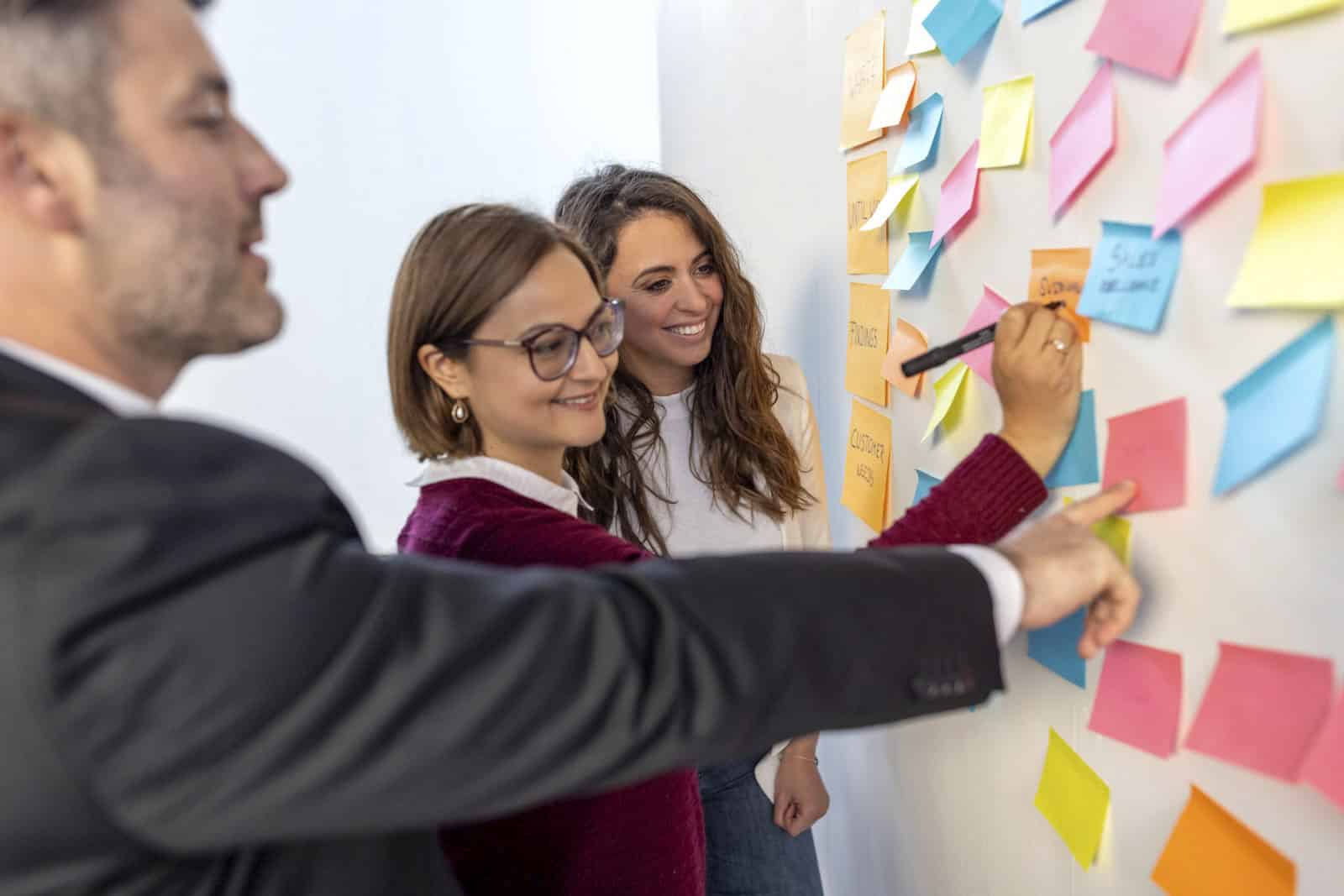 Drei Personen stehen vor einer Wand, die mit bunten Haftnotizen bedeckt ist. Eine Person schreibt mit einem Marker auf eine Notiz, während die anderen beiden auf verschiedene Notizen zeigen und lächeln. Sie scheinen an einer gemeinsamen Brainstorming-Sitzung teilzunehmen. © Fotografie Tomas Rodriguez
