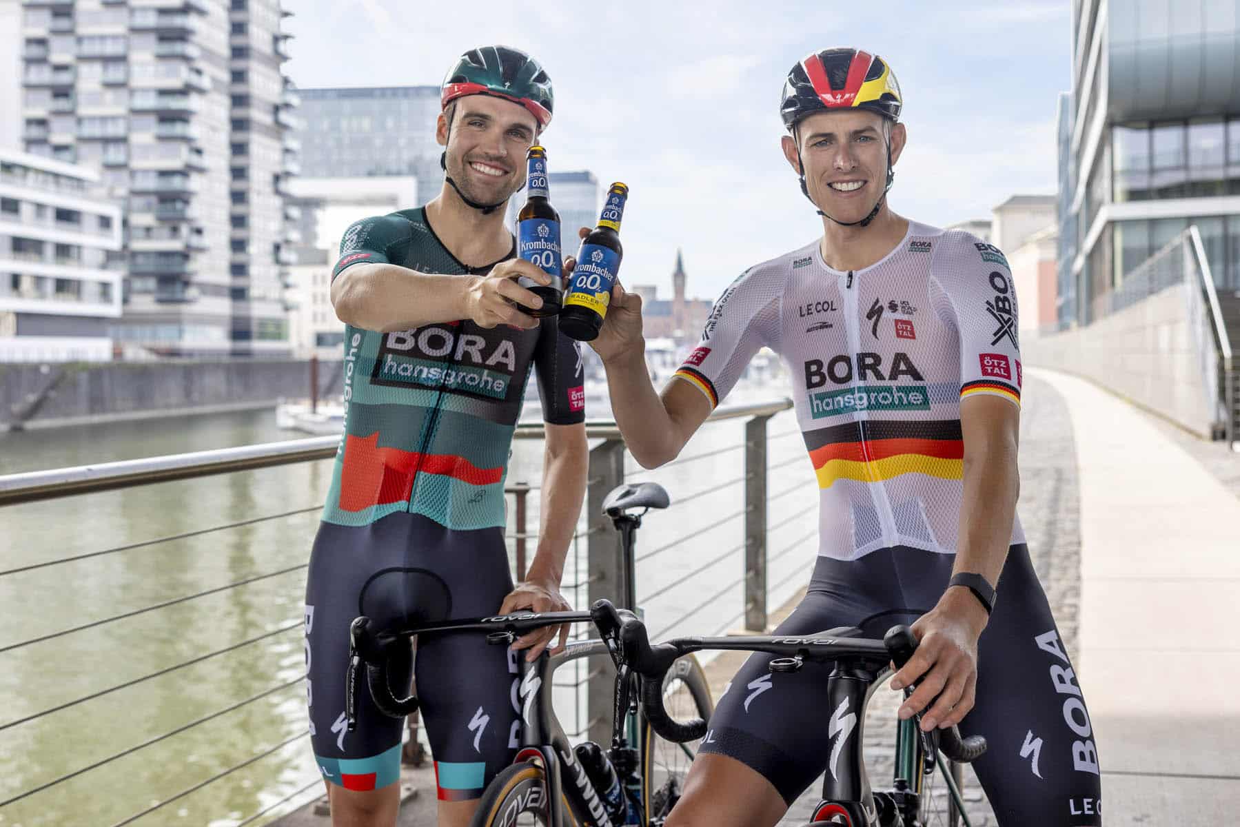 Zwei Radfahrer des Teams BORA - hansgrohe in Team-Trikots stoßen mit alkoholfreiem Bier an einem Fluss mit modernen Gebäuden im Hintergrund an. Sie halten ihre Fahrräder und lächeln in die Kamera. © Fotografie Tomas Rodriguez