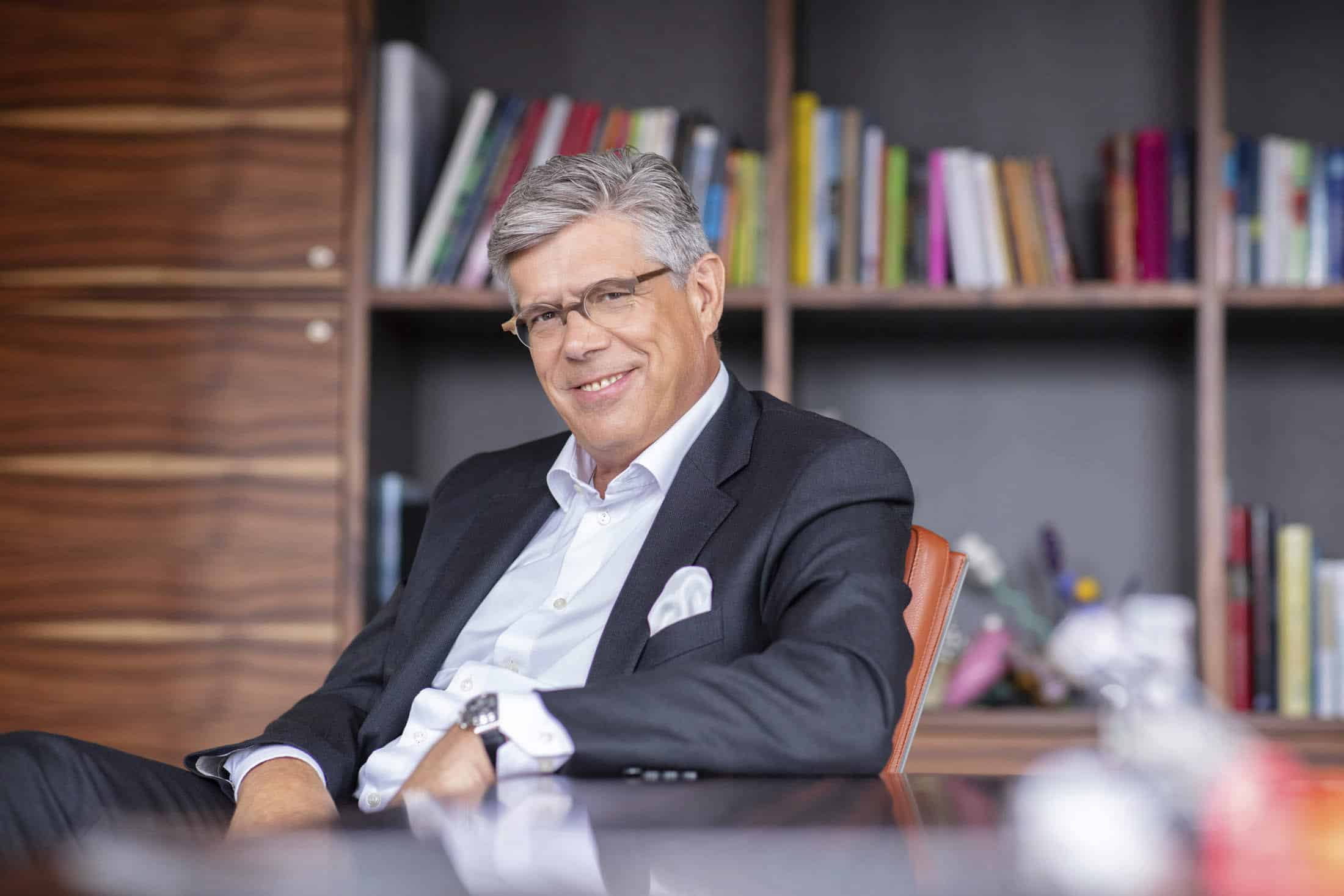 Ein lächelnder Mann mit grauem Haar, Brille und schwarzem Anzug mit weißem Hemd sitzt auf einem Bürostuhl. Im Hintergrund steht ein Holzregal mit Büchern und verschiedenen Gegenständen. © Fotografie Tomas Rodriguez
