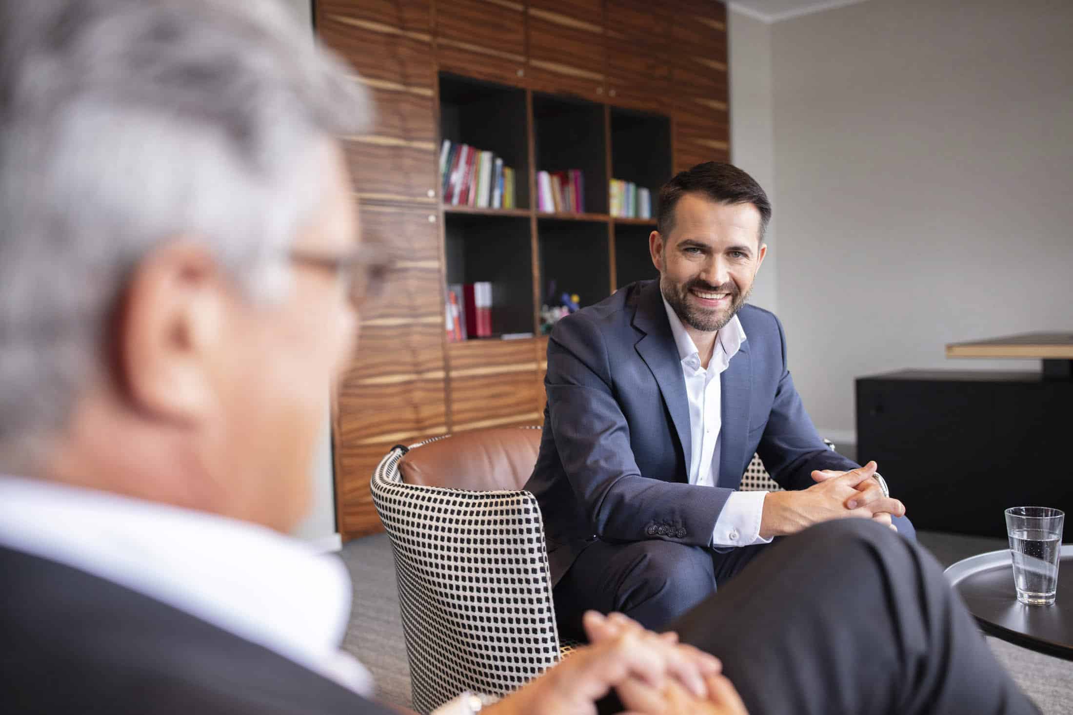 Zwei Männer in Anzügen unterhalten sich in einem Büro. Ein Mann mit grauem Haar ist von hinten zu sehen, während der andere mit Bart ihm gegenübersteht und lächelt. Sie sitzen auf modernen Stühlen, im Hintergrund ist ein hölzernes Bücherregal zu sehen. © Fotografie Tomas Rodriguez