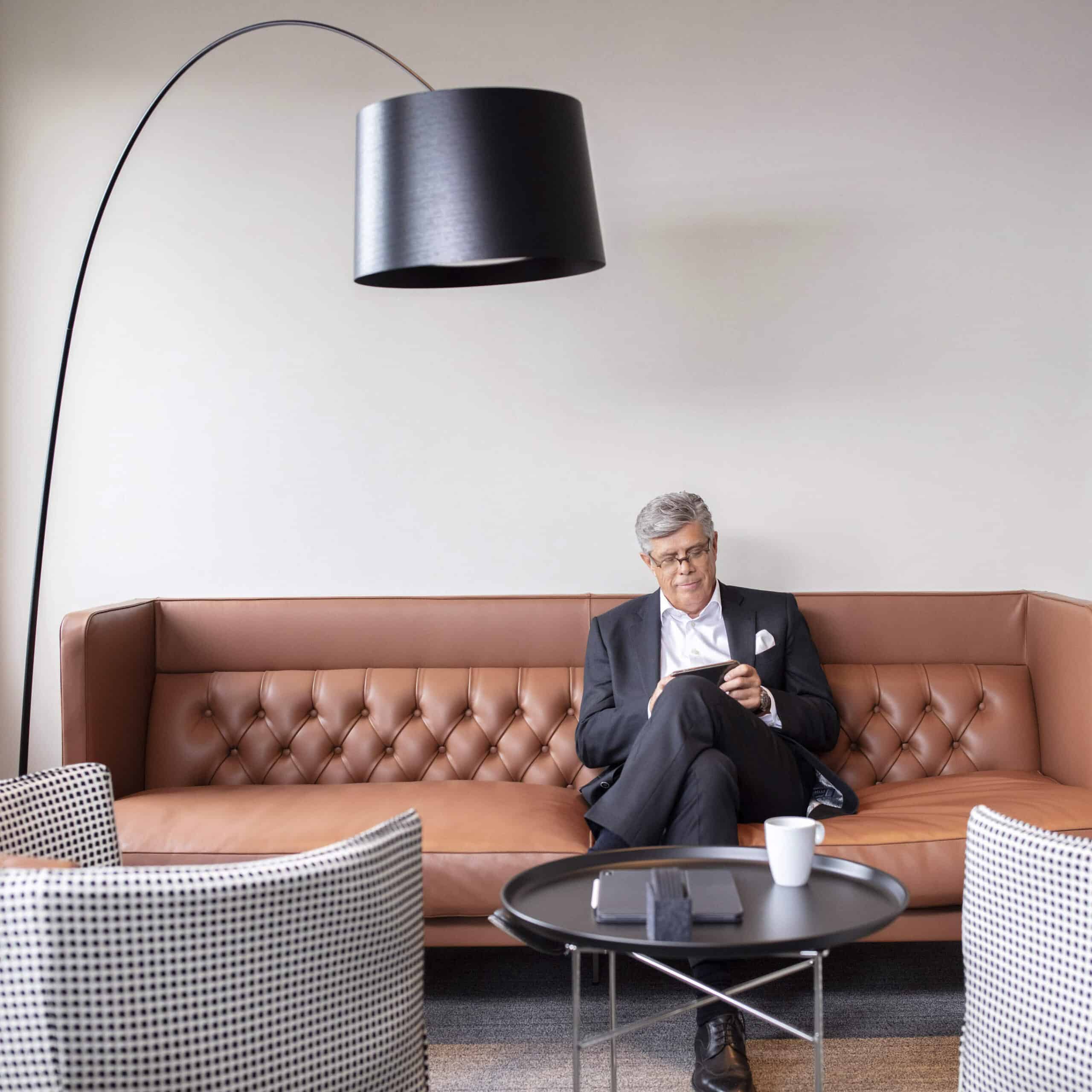 Ein Mann im Anzug sitzt auf einem gepolsterten braunen Ledersofa und schaut auf sein Telefon. Er steht unter einer schwarzen Bogenstehlampe. Im Vordergrund steht ein schwarzer runder Couchtisch mit einer weißen Tasse und einem dunklen rechteckigen Buch, daneben stehen zwei gemusterte Stühle. © Fotografie Tomas Rodriguez