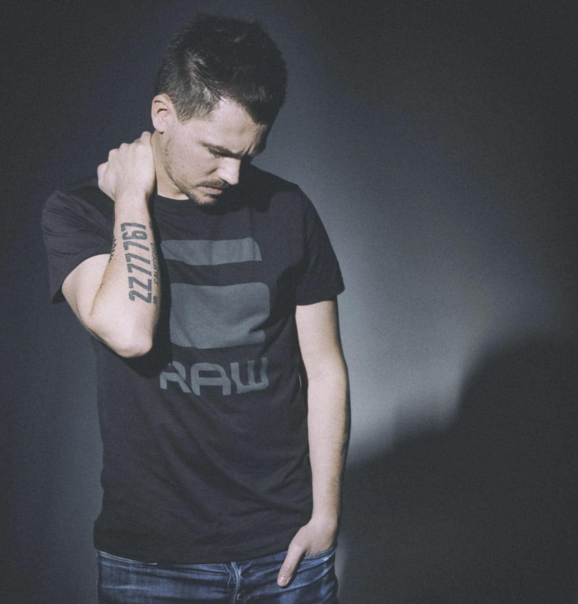 Ein Mann in einem schwarzen T-Shirt mit einem großen „RAW“-Logo steht vor einem dunklen Hintergrund. Er blickt nach unten, seine rechte Hand berührt seinen Nacken und seine linke Hand steckt in seiner Tasche. Auf seinem linken Unterarm hat er eine Tätowierung mit den Zahlen „22776“. © Fotografie Tomas Rodriguez