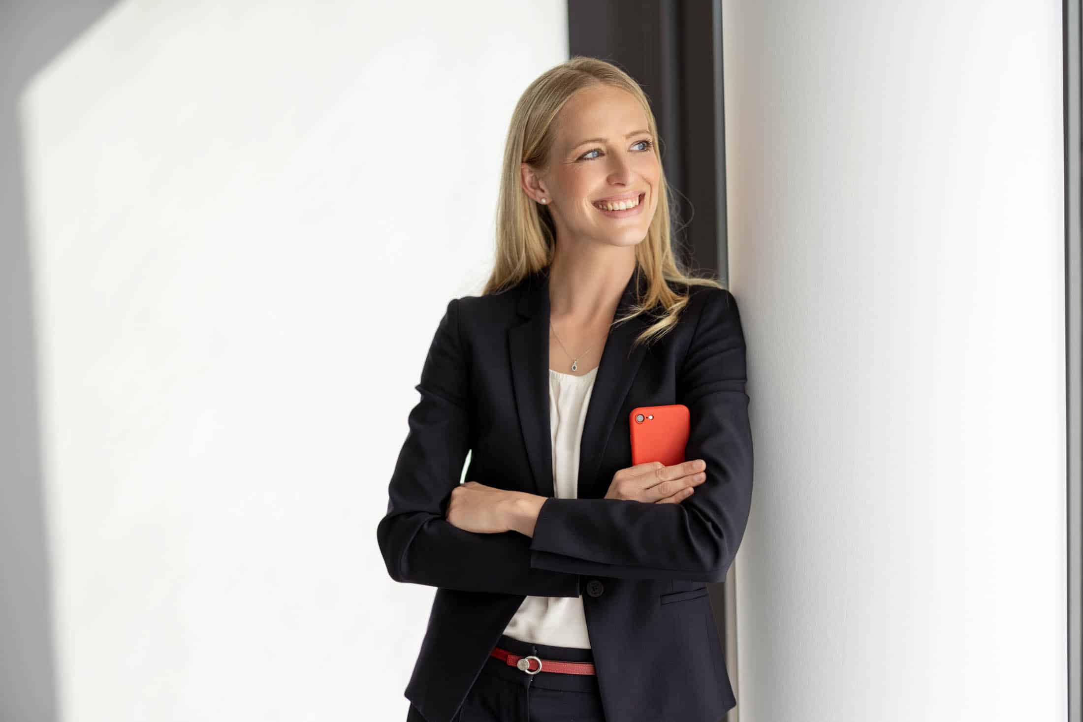 Eine Frau im Businessanzug steht lächelnd vor einer weißen Wand und hält mit verschränkten Armen ein rotes Telefon in der Hand. Sie hat lange blonde Haare und schaut zur Seite. Der Hintergrund ist hell und minimalistisch. © Fotografie Tomas Rodriguez