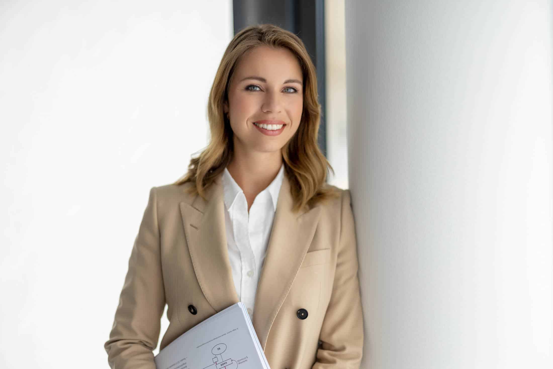 Eine Frau mit langen Haaren in einem beigen Blazer und einem weißen Hemd lehnt lächelnd an einer weißen Wand. Sie hält ein Notizbuch mit einem Diagramm auf dem Umschlag und steht in einer gut beleuchteten, modernen Umgebung. © Fotografie Tomas Rodriguez