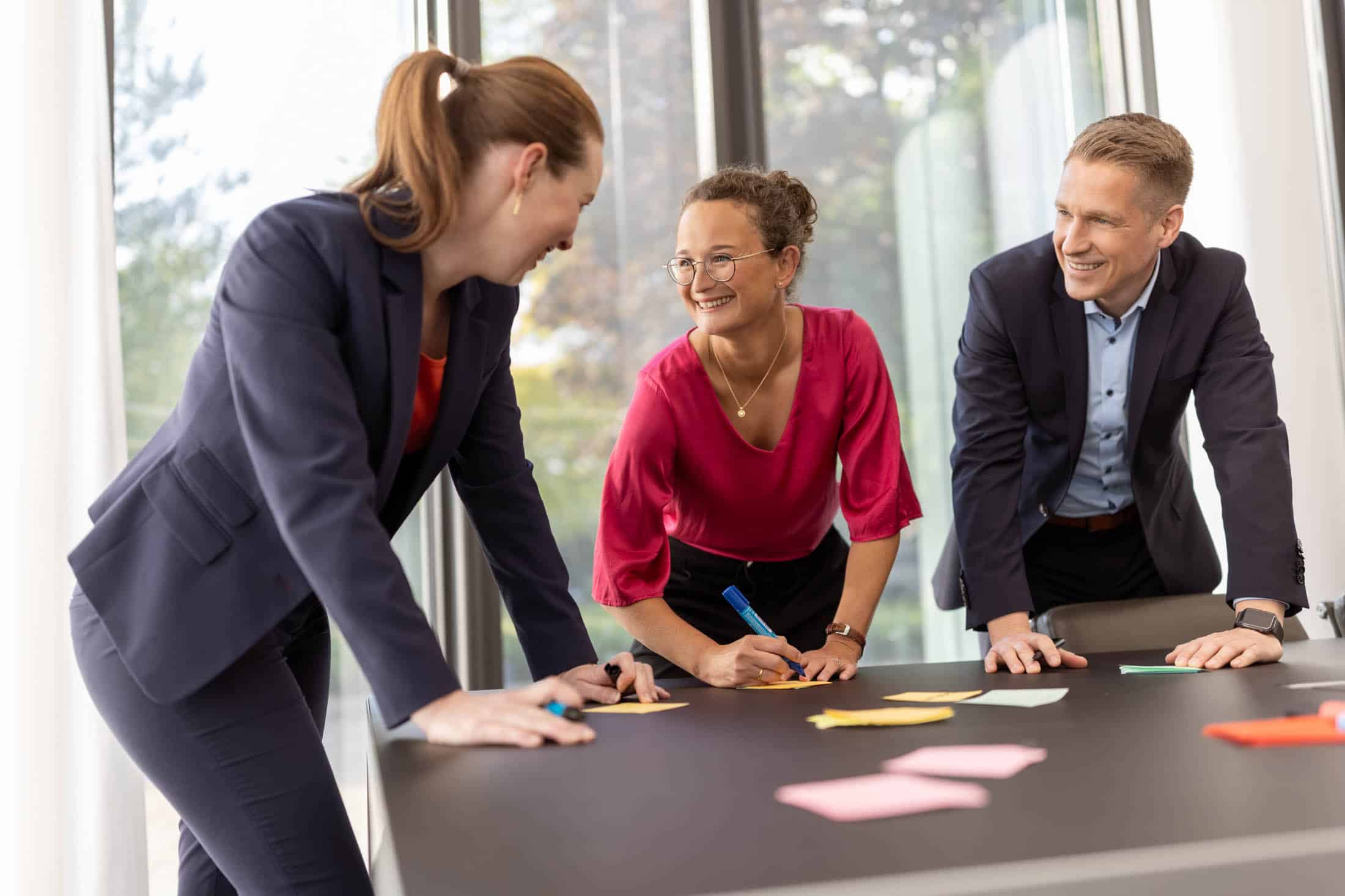 Drei Personen in Business-Kleidung sitzen lächelnd an einem Tisch und arbeiten gemeinsam mit Haftnotizen. Im Hintergrund sind große Fenster mit Blick auf Bäume zu sehen. © Fotografie Tomas Rodriguez