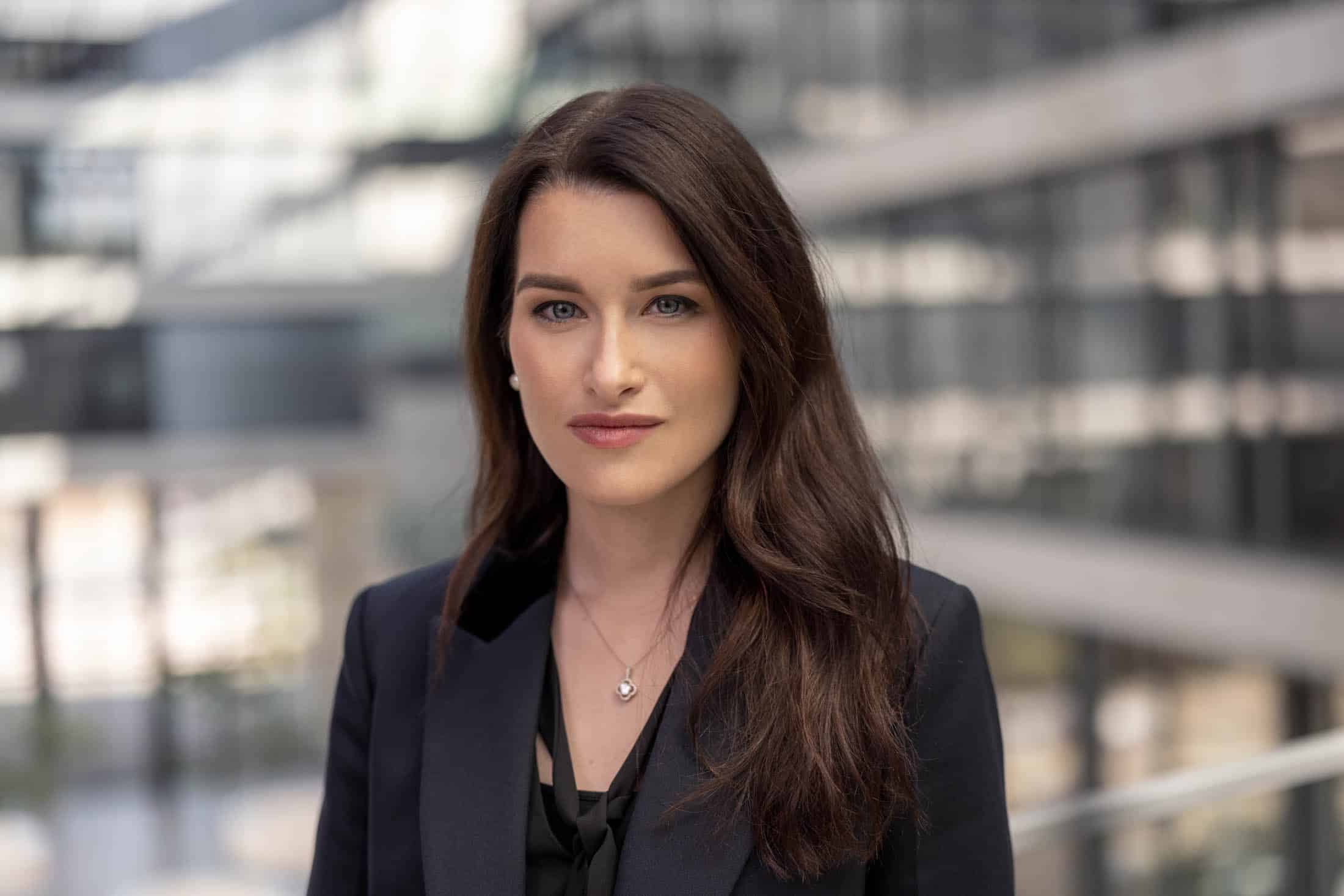 Eine Frau mit langen braunen Haaren, einem schwarzen Blazer und einer Halskette steht in einem Büro mit großen Fenstern und blickt in die Kamera. © Fotografie Tomas Rodriguez