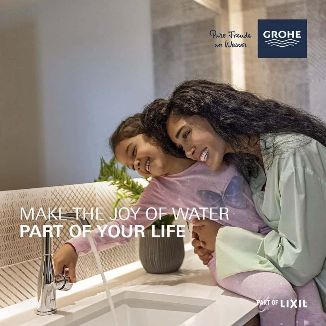 Eine Frau und ein Kind beschäftigen sich freudig mit einem Badezimmerhahn. Die Frau beugt sich über das lächelnde Kind, während beide nach dem Wasser greifen. Das Bild zeigt den Text „Machen Sie die Freude am Wasser zu einem Teil Ihres Lebens“ und das Grohe-Logo. © Fotografie Tomas Rodriguez