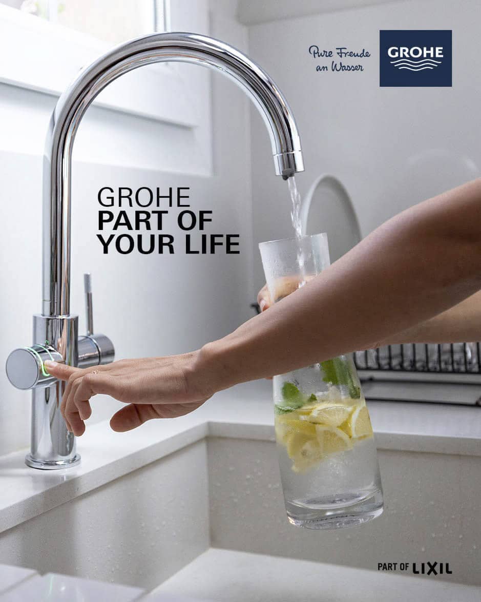 Eine Hand füllt einen durchsichtigen Krug mit Zitronen- und Limettenscheiben mit Wasser aus einem eleganten, modernen Wasserhahn. Im Hintergrund ist eine Küchenspüle neben einem Fenster zu sehen. Der Text lautet „GROHE TEIL IHRES LEBENS“. © Fotografie Tomas Rodriguez