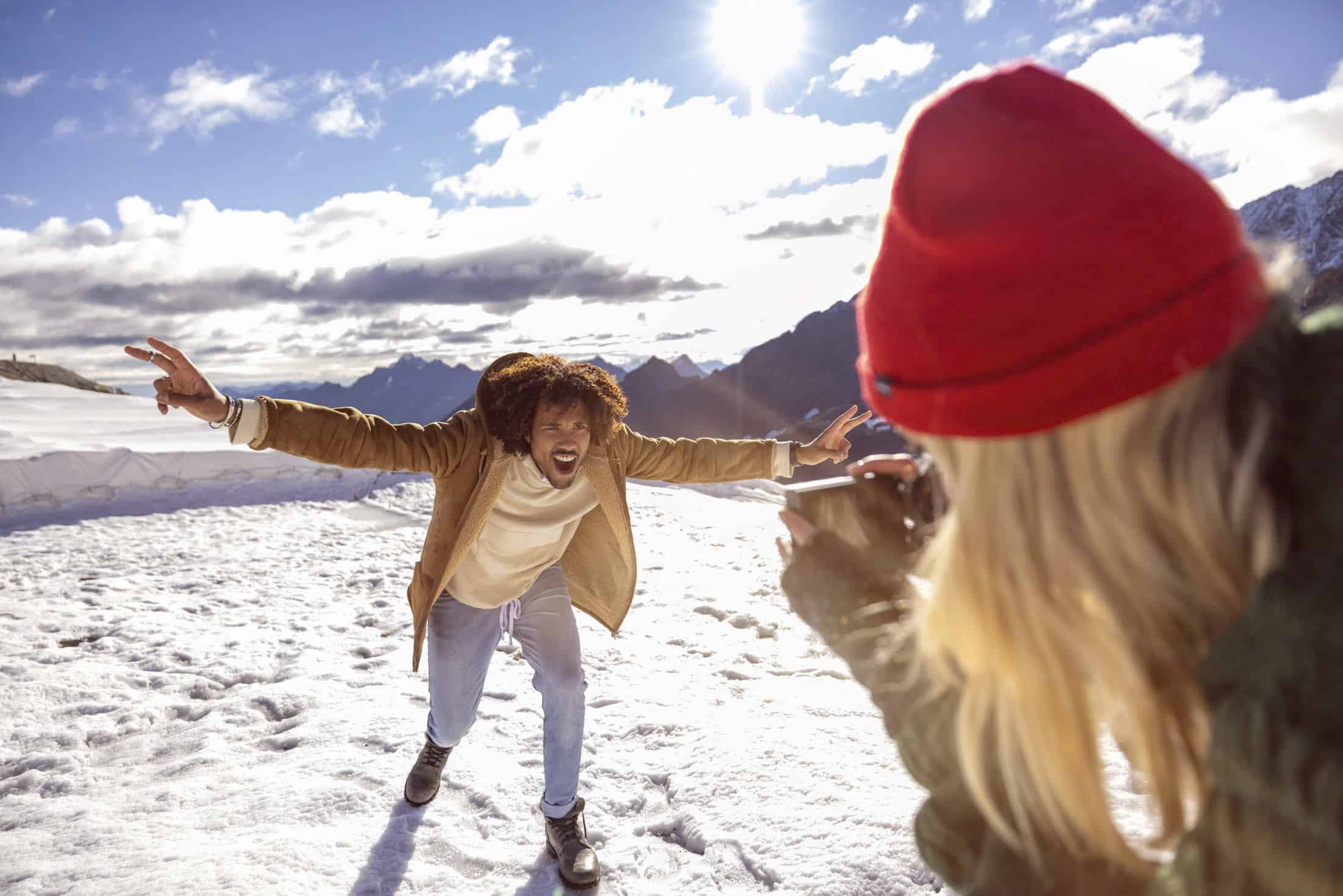 Eine Person in einem braunen Mantel und Jeans posiert fröhlich auf einem schneebedeckten Berg und streckt die Arme aus, während eine andere Person mit einer roten Mütze ein Foto von ihr macht. Die Sonne scheint hell und beleuchtet den Schnee und die entfernten Gipfel. © Fotografie Tomas Rodriguez