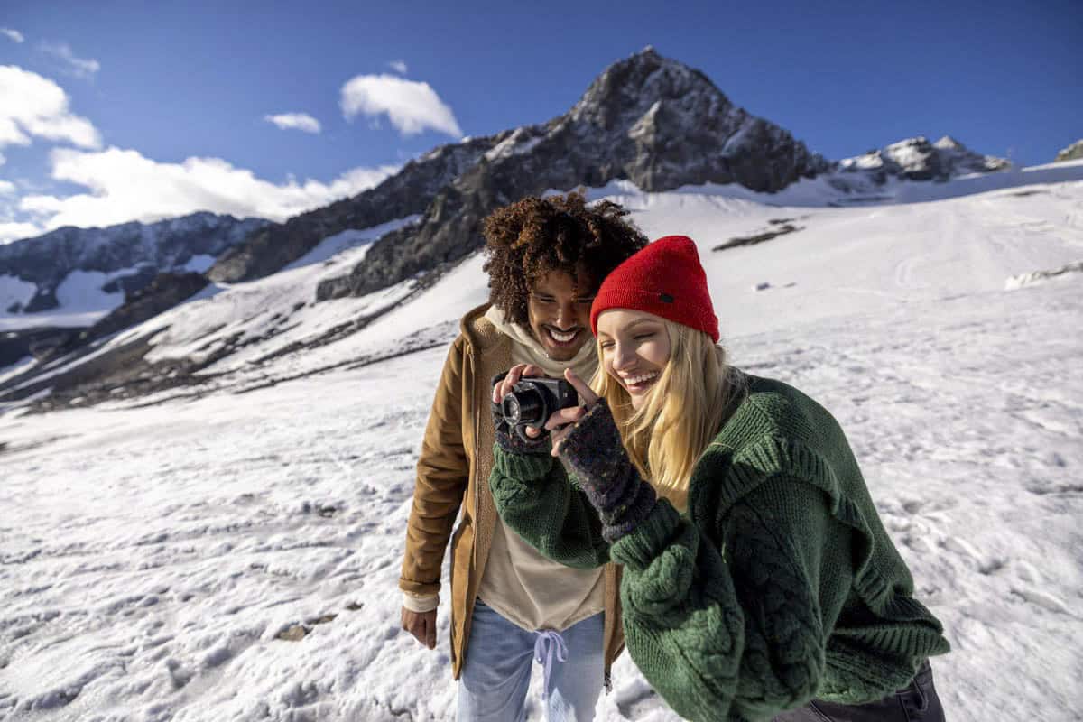 Ein Mann und eine Frau lächeln, während sie in einer verschneiten Berglandschaft in eine Kamera blicken. Die Frau trägt eine rote Mütze und einen grünen Pullover, und der Mann trägt eine braune Jacke. Der Himmel ist klar mit ein paar Wolken. © Fotografie Tomas Rodriguez