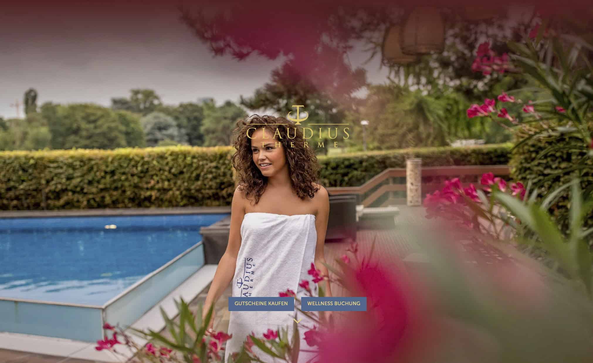 In ein Handtuch gehüllte Frau spaziert an einem Pool entlang, der von Grün und rosa Blumen umgeben ist. Die Szene vermittelt einen ruhigen Eindruck eines Spas im Freien, wobei ein Logo und Navigationsoptionen über das Bild gelegt sind. © Fotografie Tomas Rodriguez