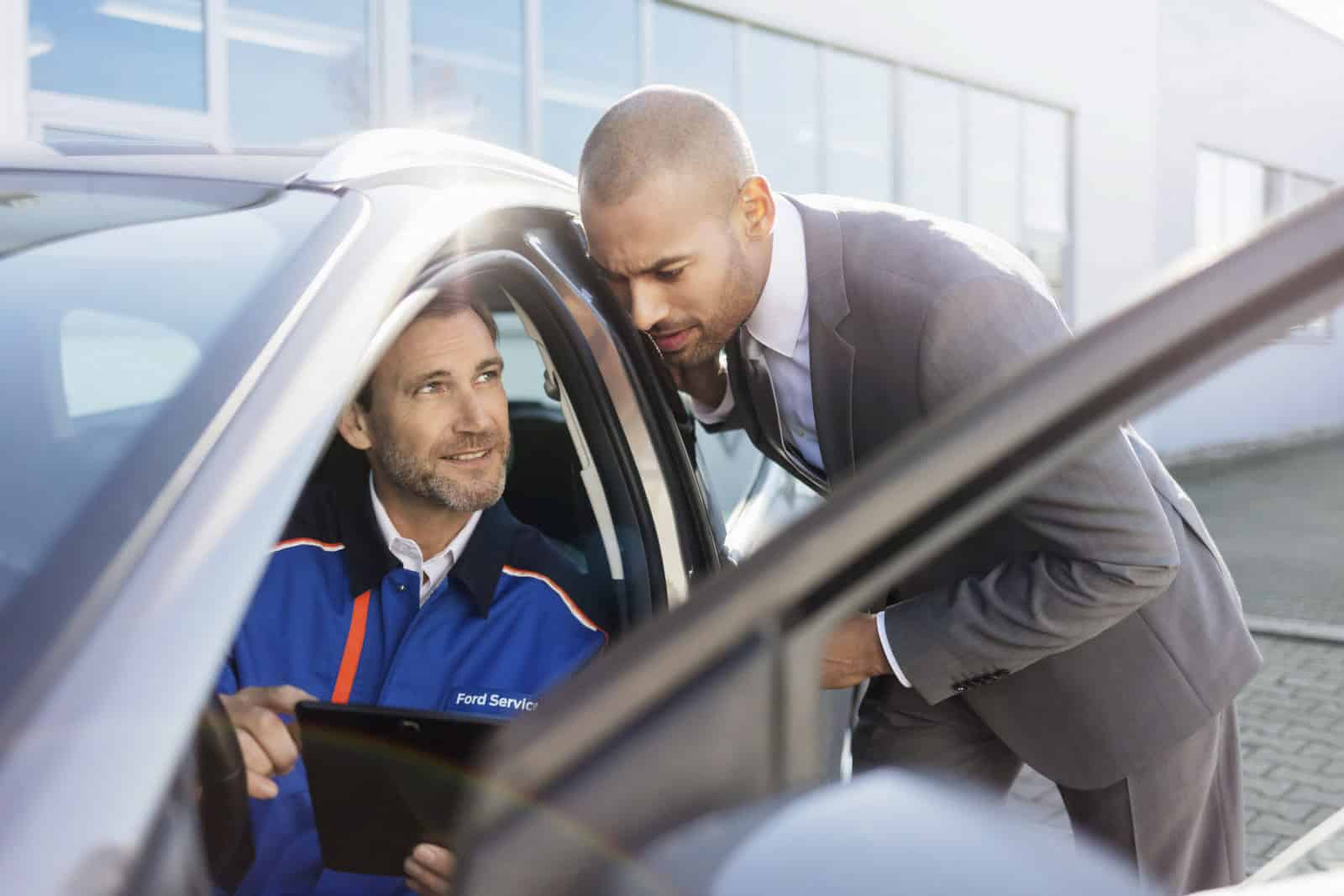 Ein Mann in einer blauen Ford-Servicejacke sitzt in einem Auto und zeigt einem anderen Mann im Anzug, der sich durch die offene Autotür hineinlehnt, ein Tablet. Sie scheinen etwas zu besprechen, das mit dem Fahrzeug zu tun hat. © Fotografie Tomas Rodriguez