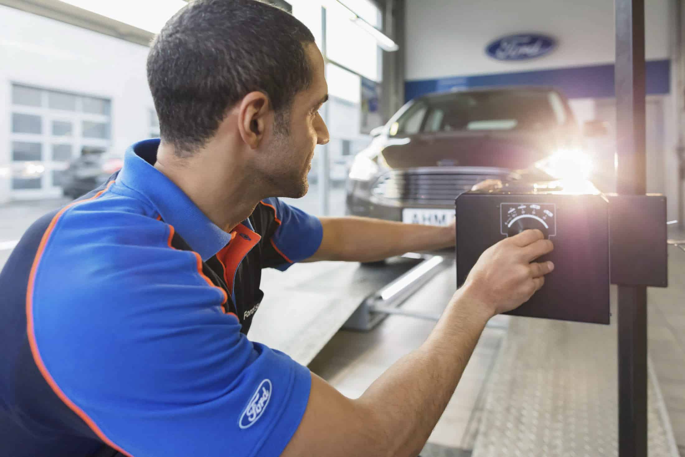 Ein Mechaniker in blau-schwarzer Uniform richtet in einer Werkstatt eine auf die Scheinwerfer eines Autos gerichtete Ausrüstung aus. Auf seinem Ärmel ist ein Ford-Logo zu sehen. Das Fahrzeug ist auf einer Plattform geparkt, im Hintergrund sind Garagentore zu sehen. © Fotografie Tomas Rodriguez