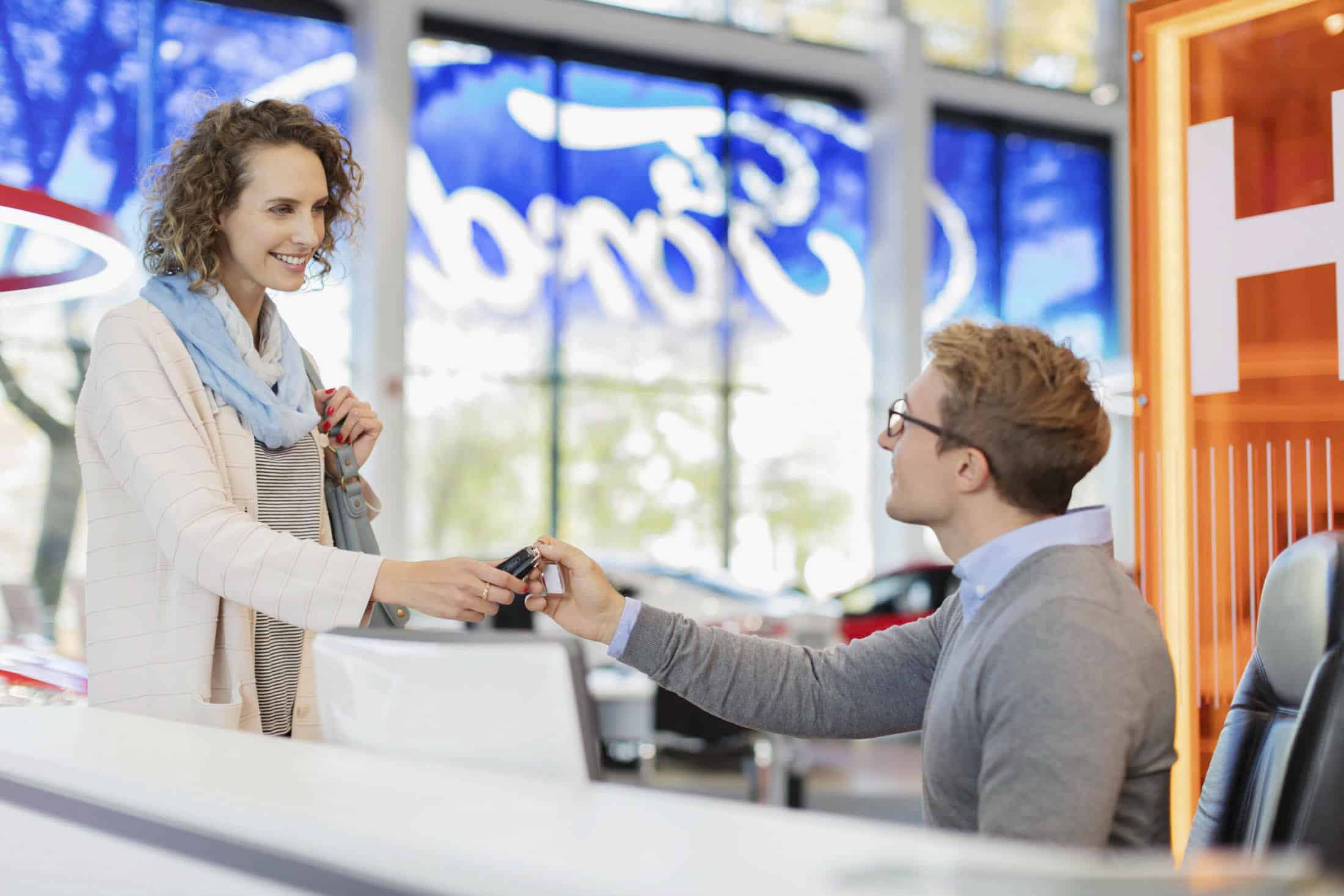 Eine Frau gibt einem Mann, der in einem hell erleuchteten Büro mit großen Fenstern hinter einem Schreibtisch sitzt, einen Autoschlüssel. Beide lächeln. Im Hintergrund ist ein großes blaues Logo für „Ford“ zu sehen. © Fotografie Tomas Rodriguez