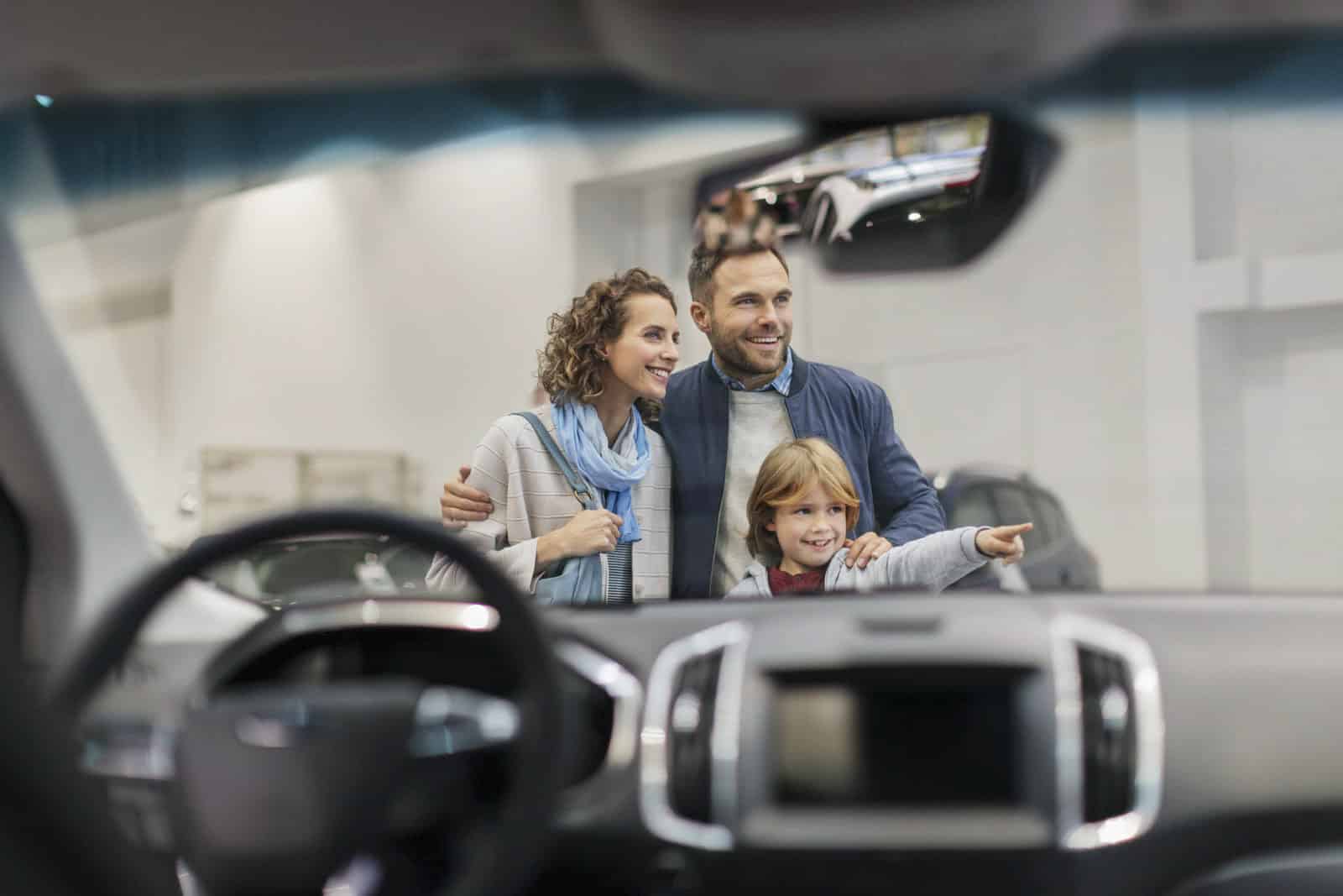 Eine dreiköpfige Familie – zwei Erwachsene und ein Kind – steht in einem Autohaus, gesehen durch die Windschutzscheibe eines Autos. Das Kind zeigt aufgeregt nach vorne, während die Erwachsenen lächeln und der Vater sanft die Schulter des Kindes hält. © Fotografie Tomas Rodriguez