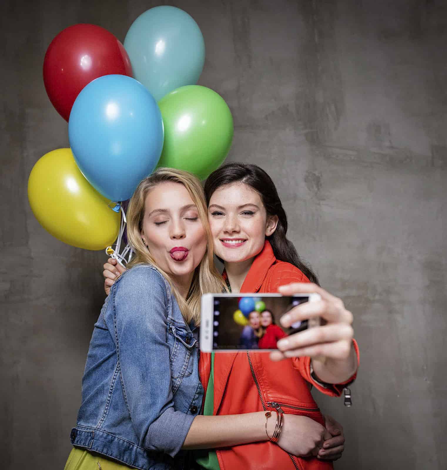 Zwei Frauen machen ein Selfie. Eine hält bunte Luftballons und beide lächeln. Die Frau links streckt verspielt die Zunge heraus. Der Hintergrund ist eine schlichte, graue Wand. © Fotografie Tomas Rodriguez