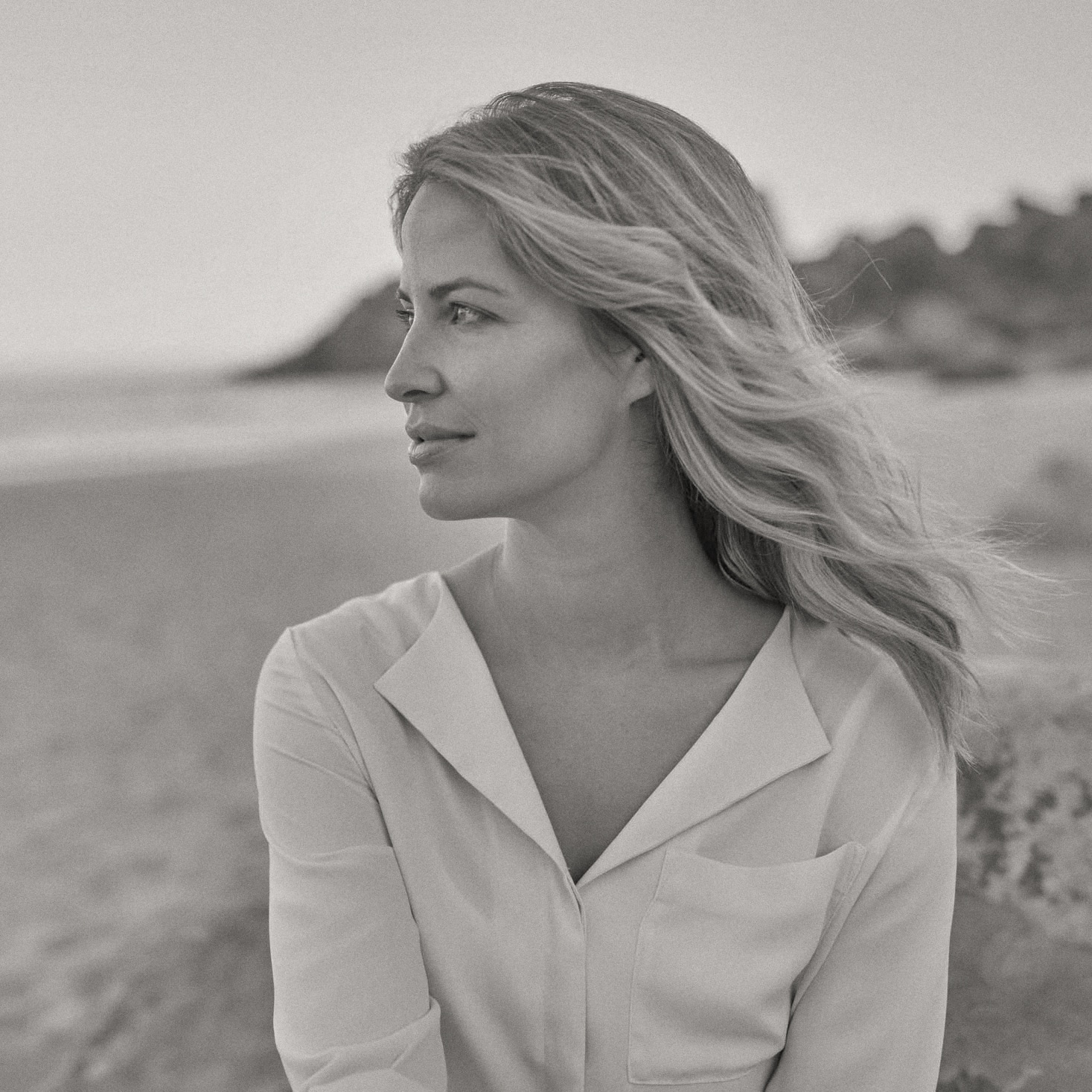 Eine Frau mit langen Haaren blickt nachdenklich zur Seite, während sie auf einem Felsen am Strand sitzt. Sie trägt eine helle, langärmelige Bluse. Die Umgebung ist ruhig, mit einem verschwommenen Hintergrund aus Sand und entfernten Felsen. Das Bild ist in Schwarzweiß. © Fotografie Tomas Rodriguez