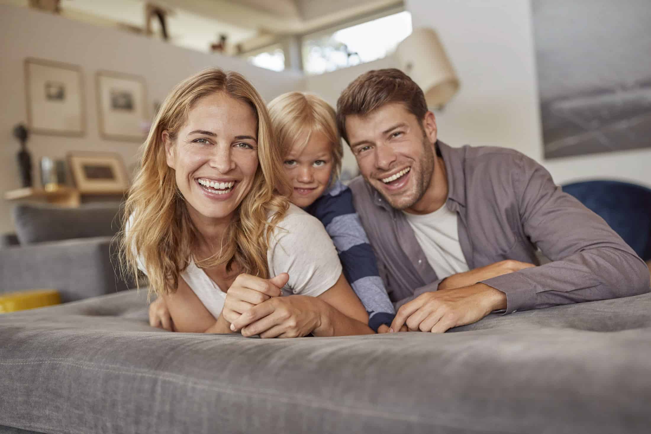 Eine glückliche dreiköpfige Familie, bestehend aus einer Frau, einem Mann und einem Kind, liegt auf einem grauen Sofa in einem hellen Wohnzimmer. Sie lächeln und schauen in die Kamera. Der Raum ist modern eingerichtet, mit Kunstwerken an den Wänden und sanfter Beleuchtung. © Fotografie Tomas Rodriguez