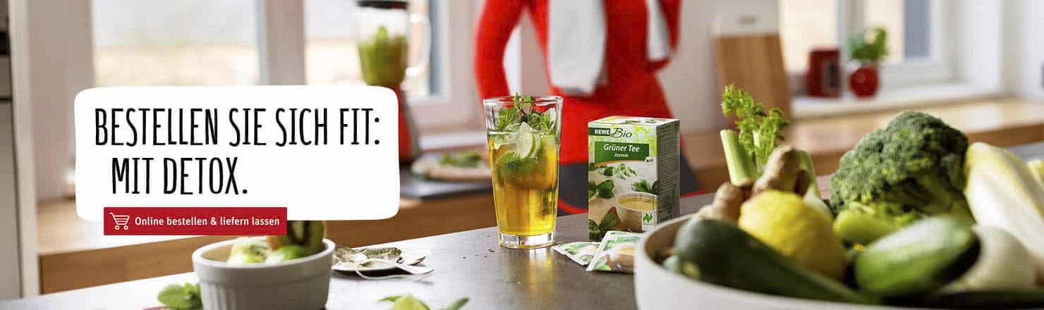 Eine Küchentheke mit einem Glas Eistee, Obst und Gemüse, darunter Brokkoli und Zucchini. Eine Schachtel Kräutertee zum Entgiften steht darauf. Der deutsche Text auf der linken Seite lautet: „Bestellen Sie sich fit: mit Detox“ und lädt dazu ein, Entgiftungsprodukte online zu bestellen. © Fotografie Tomas Rodriguez