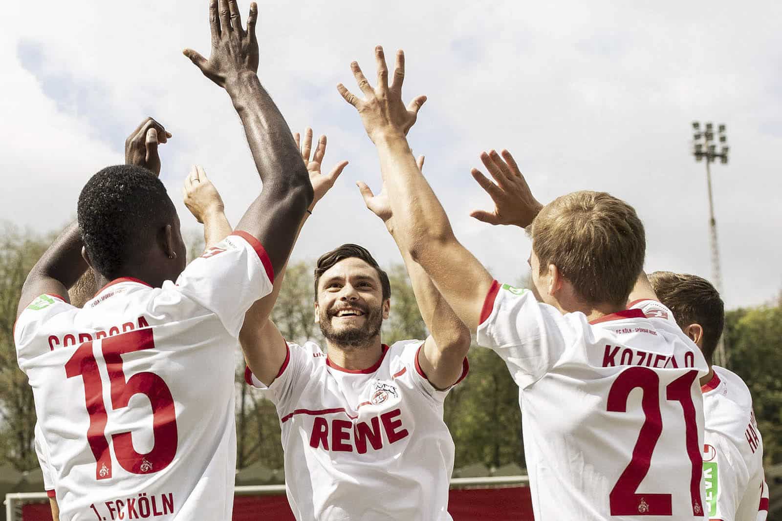 Fünf Fußballspieler in weißen Trikots mit roten Akzenten geben sich High Fives und feiern auf einem Außenfeld. Die Rücken der Spieler sind größtenteils sichtbar, so dass auf zwei Trikots die Nummern 15 und 21 zu sehen sind. Im Hintergrund sind ein Flutlicht und Bäume zu sehen. © Fotografie Tomas Rodriguez