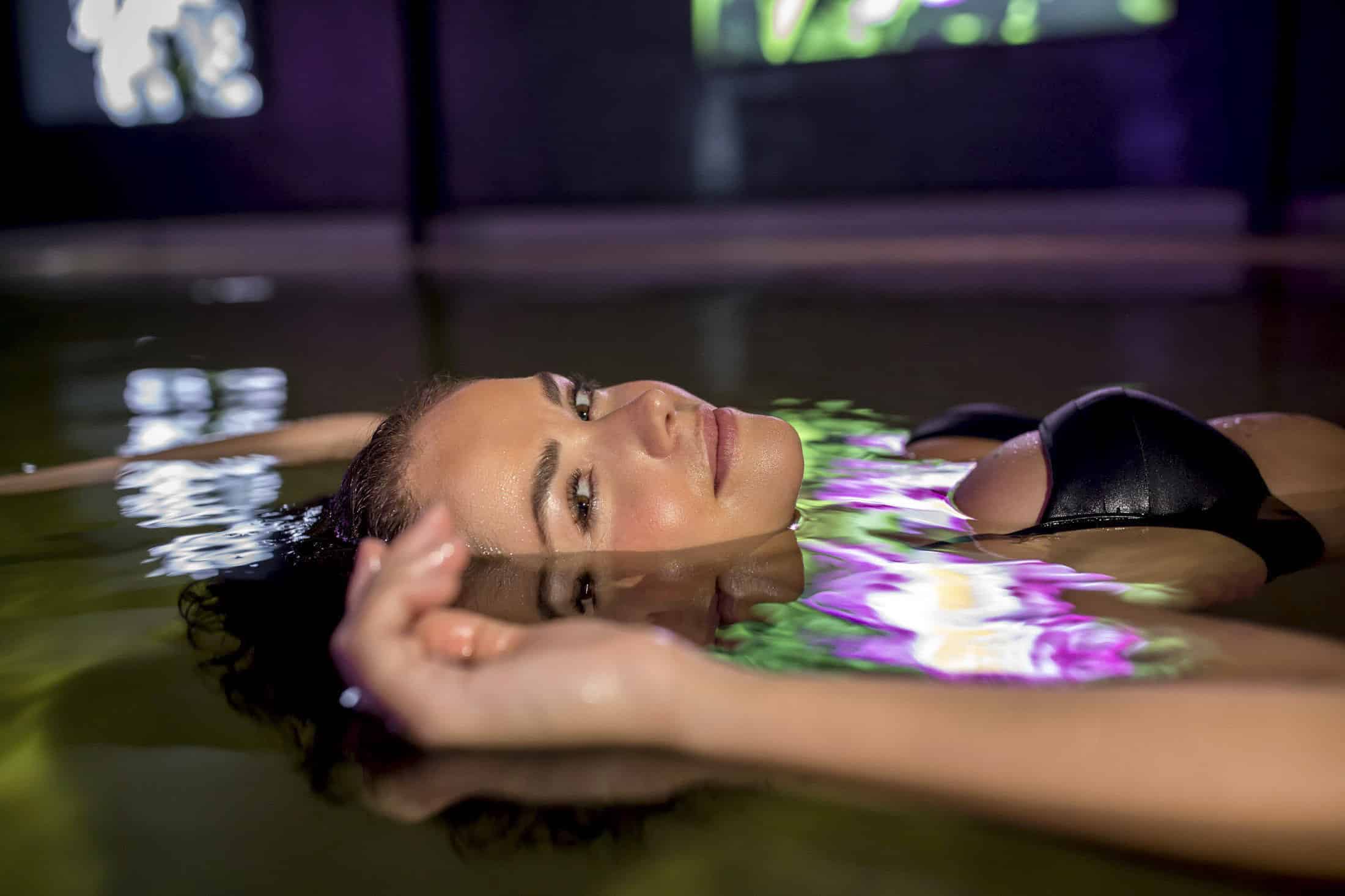Eine Person, die in einem schwarzen Badeanzug auf dem Rücken in einem Pool schwimmt, mit heiterem Gesichtsausdruck. Das Wasser reflektiert bunte Lichter und erzeugt ein lebendiges, künstlerisches Muster. Sanftes Licht verstärkt die friedliche und entspannte Atmosphäre. © Fotografie Tomas Rodriguez